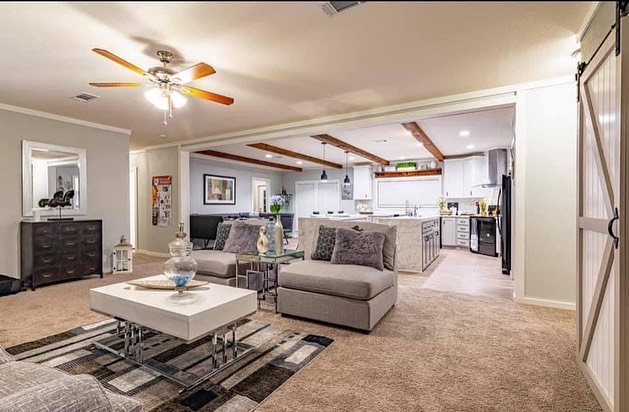 A modern open-concept living room and kitchen with neutral tones. The living area features comfy gray seating, a ceiling fan, carpet, and a patterned rug, while the kitchen showcases white cabinets, wood ceiling beams, and bright lighting, creating an inviting ambiance.