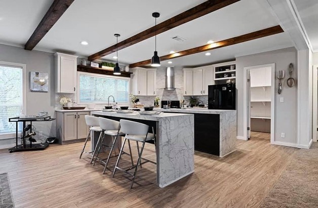 Spacious modern kitchen with light wood flooring and white cabinets. Features a large marble island with bar stools, black appliances, and exposed wooden beams.
