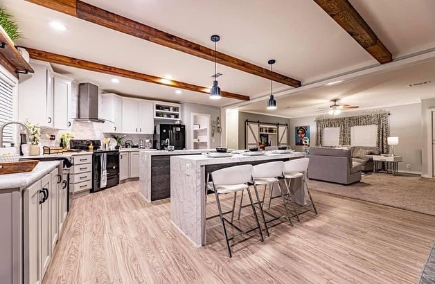 A modern open-concept kitchen with a living area features light wood floors, a large island and white cabinets. Exposed beams add a rustic touch.