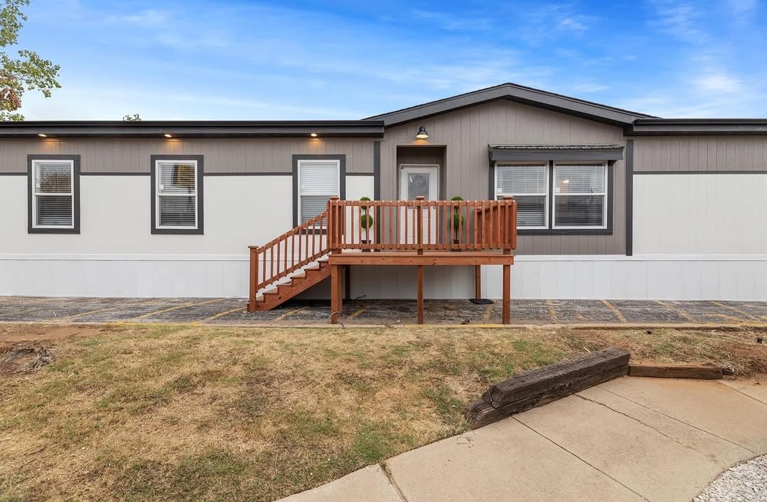 A modern mobile home with a gray and white exterior, featuring a small wooden porch and steps. The lawn is slightly dry under a bright blue sky.