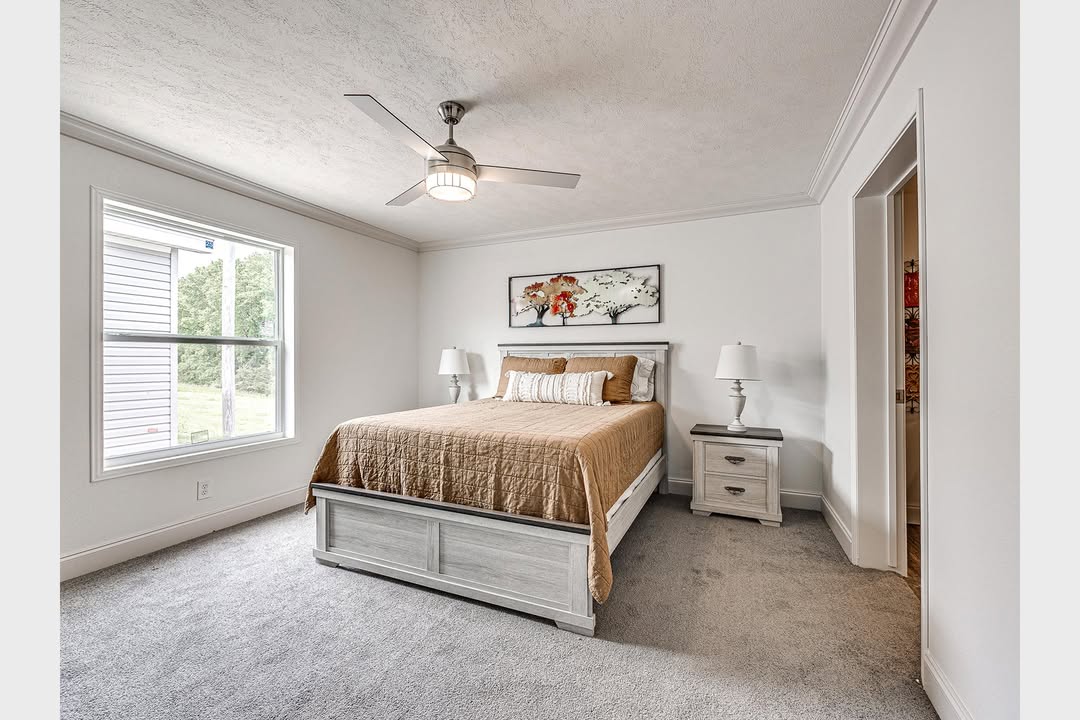 A cozy bedroom with a beige bedspread on a double bed, flanked by two white nightstands and lamps. A floral artwork hangs above. Soft natural light streams from a window, and a ceiling fan is centered overhead.