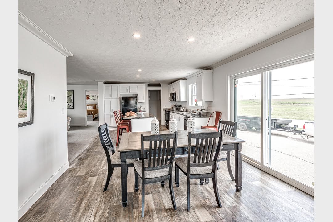 Dining area with a dark wooden table and six chairs on a wooden floor. Bright kitchen in background with white cabinets and a large sliding glass door showing an outdoor scene. Cozy and spacious feel.