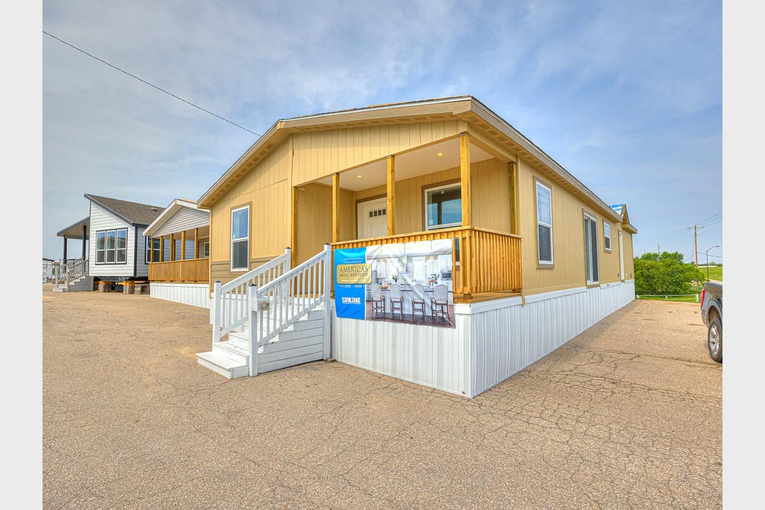 Single-story manufactured home with beige siding and white trim, featuring a covered porch and banner. The setting is sunny, with a clear blue sky.