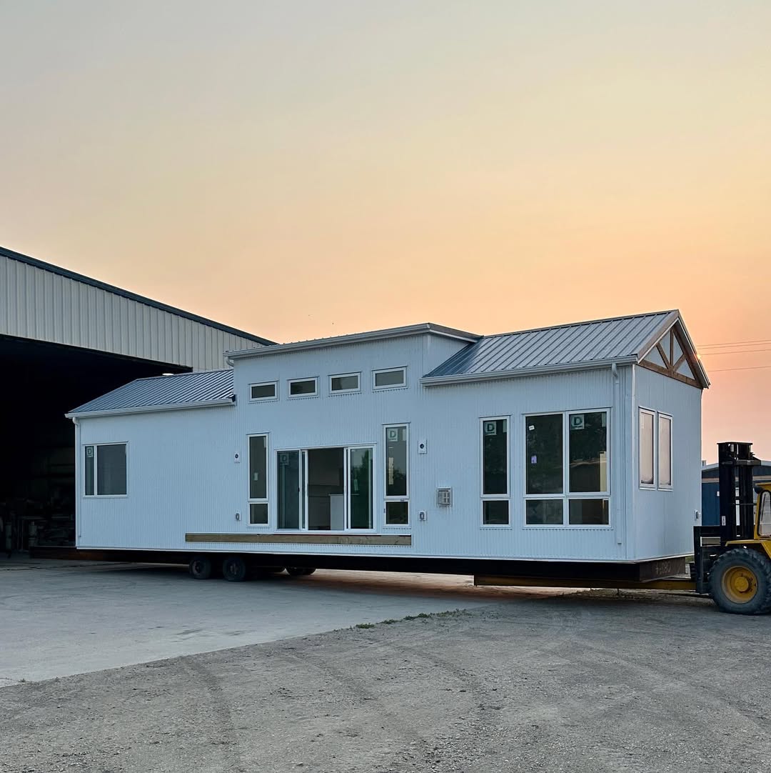 A modern white tiny house with several windows is being transported by a forklift. It sits on wheels outside a large storage building under a soft orange sky.