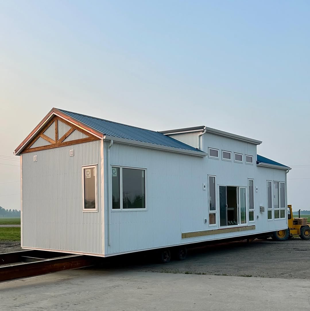 A modern tiny house on wheels with white corrugated walls and blue roof panels. It is situated on a flatbed trailer in an open area, conveying mobility.