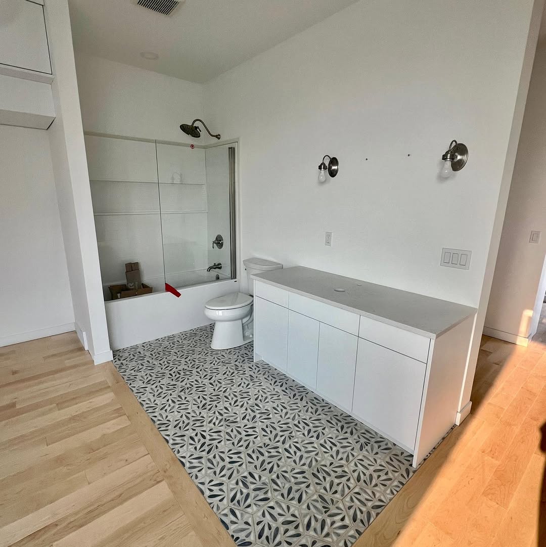 Modern bathroom with geometric patterned tile floor, white walls, and minimalistic fixtures. Bathtub with glass door and white counter evoke a clean, serene feel.