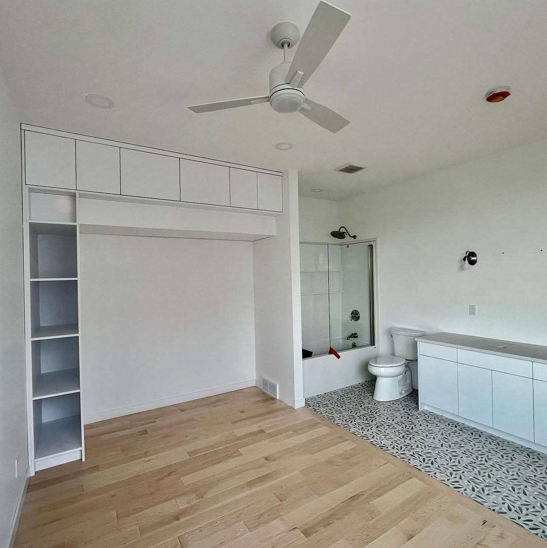 Minimalist room with white walls, wooden floor, and a ceiling fan. Features built-in shelves, a bathroom with a tub, and patterned tile flooring.