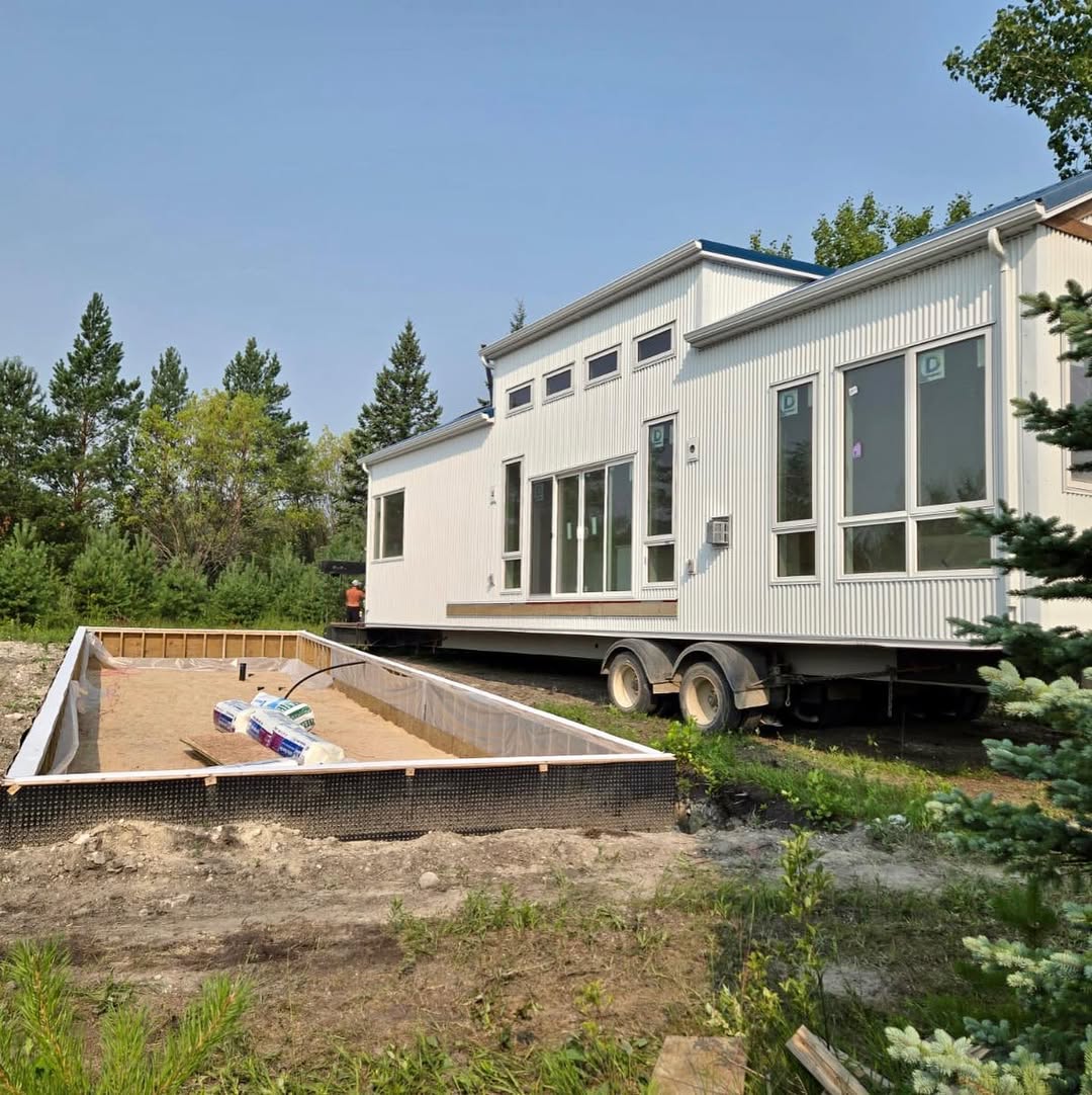 Modern, single-story white modular home on wheels, set in a lush, wooded area. Nearby, a rectangular foundation is being prepared, with bags of materials visible.