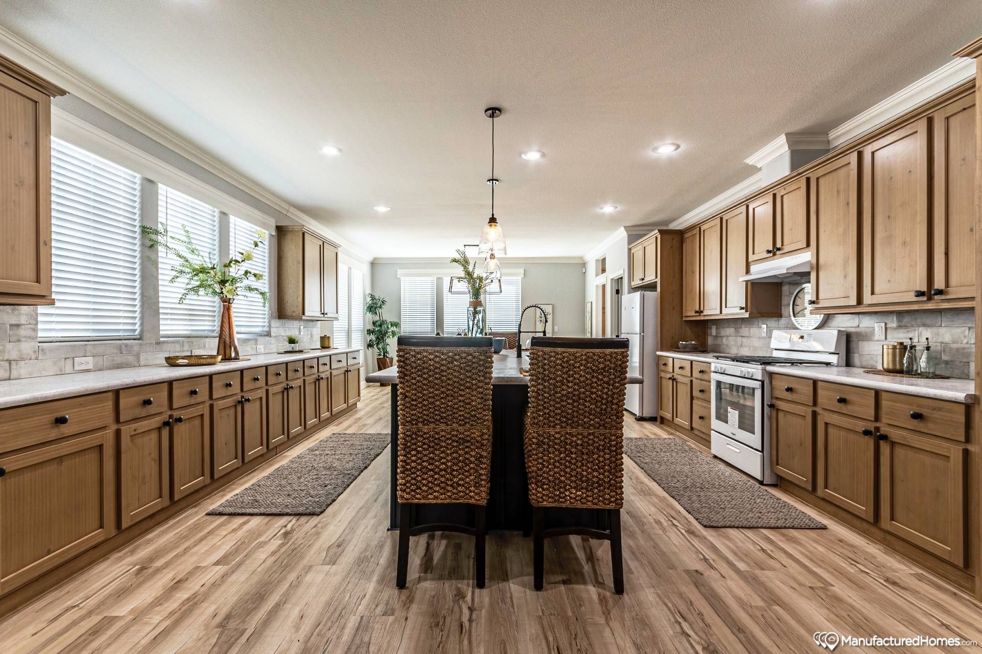 Spacious kitchen with warm wood cabinets and flooring, featuring a large island with wicker chairs. Bright lighting and plant decor add a cozy feel.
