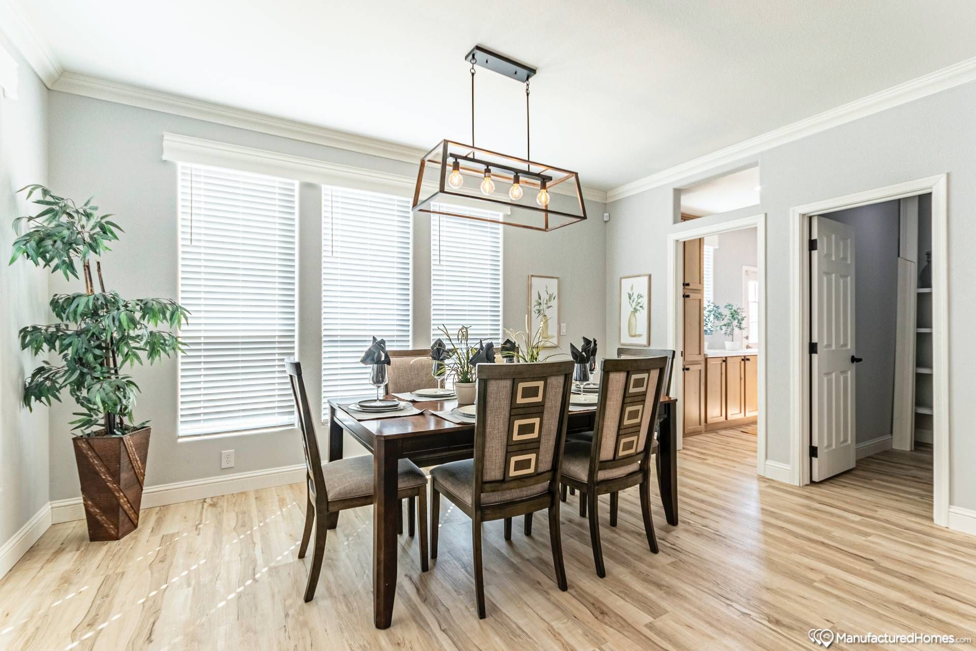 Bright dining room with a large wooden table set for six under a modern chandelier. Light streams through tall windows, highlighting sleek wood floors.