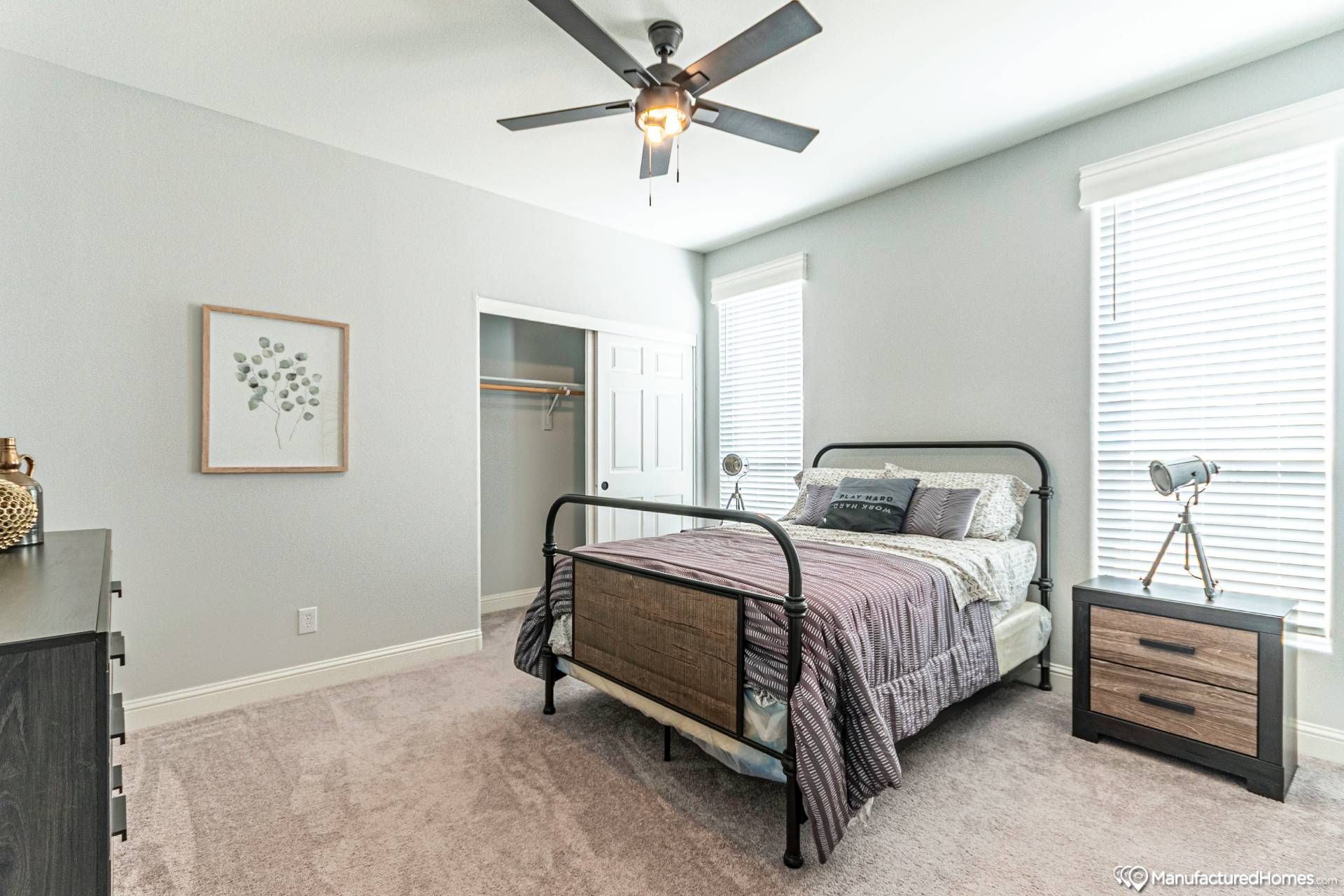 Modern bedroom with a metal-framed bed, gray bedding, and two large windows. A ceiling fan hangs above, and a small wooden dresser and open closet are visible.