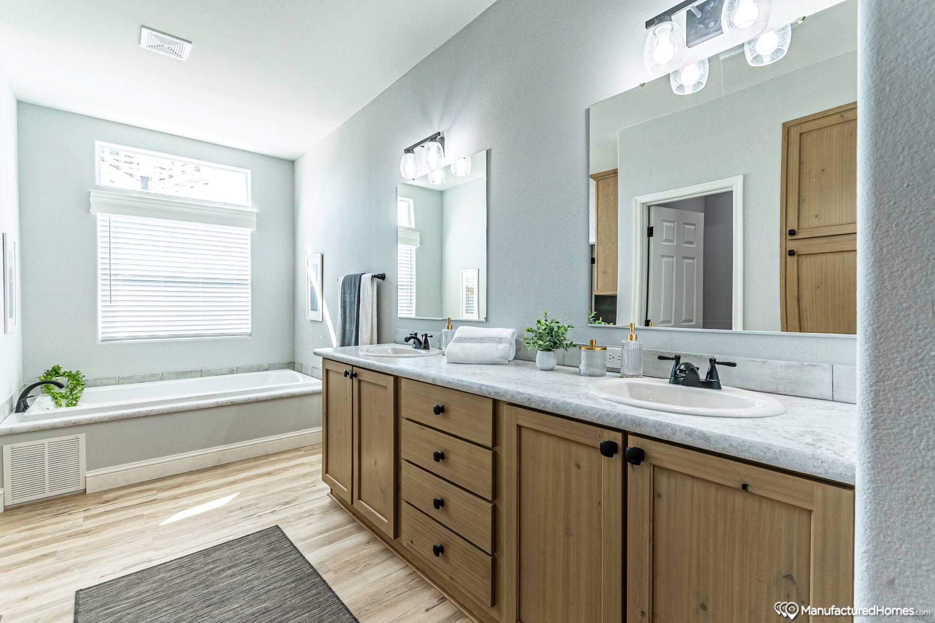 Bright, modern bathroom with dual sinks on a wooden vanity, large mirror, and bathtub under a window. Light walls and flooring enhance spacious feel.