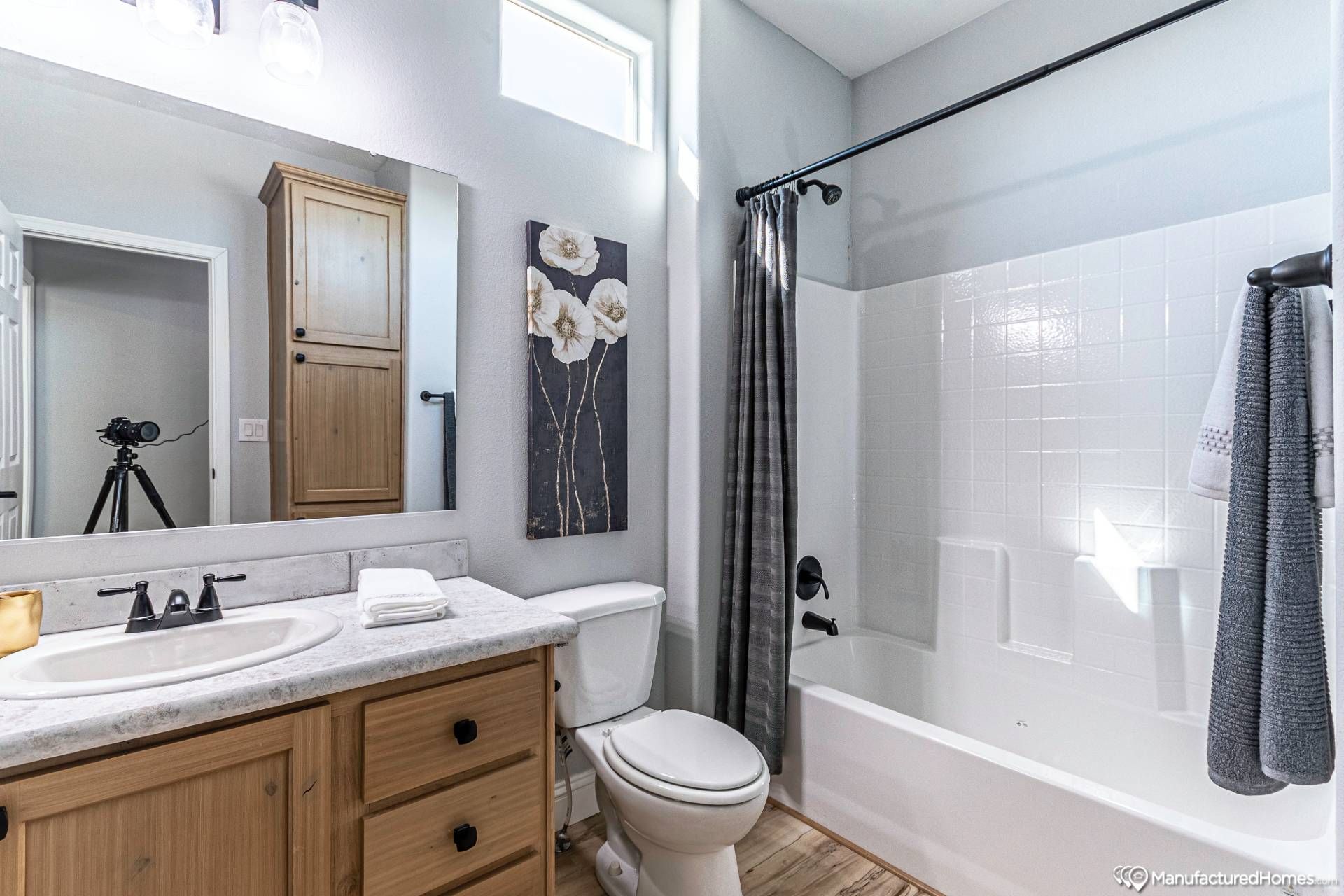 Bright bathroom featuring a wooden vanity with marble countertop, black fixtures, a white bathtub with a gray curtain, and floral wall art. Clean and modern.