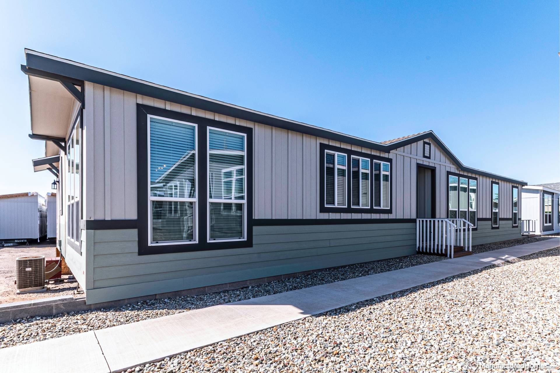 Modern manufactured home in bright sunlight, featuring a beige exterior with dark trim. A gravel path runs alongside, under a clear blue sky.