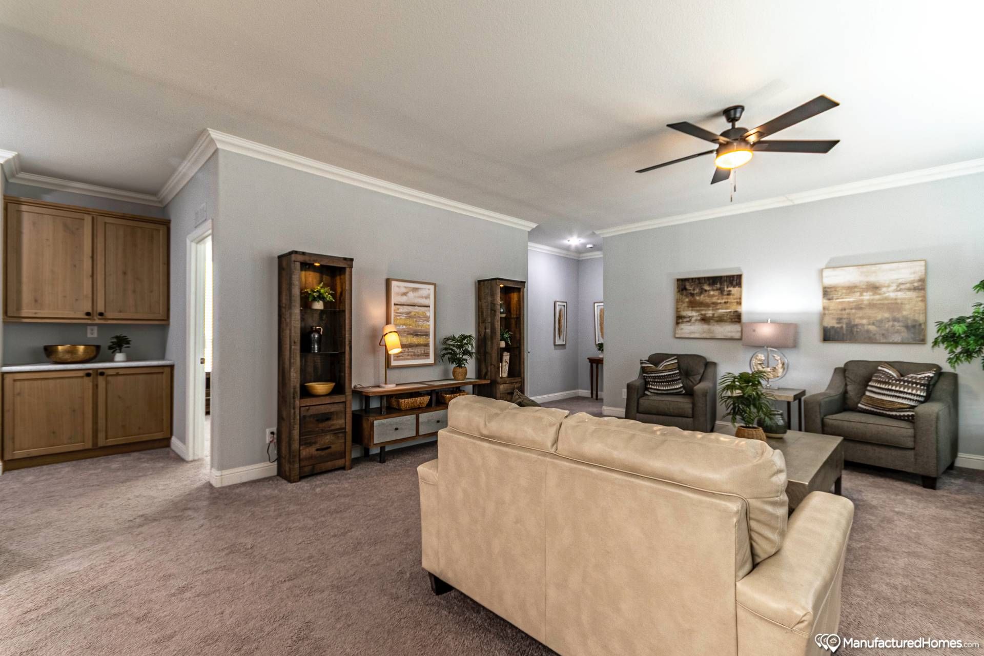 Spacious living room with beige sofa, two armchairs, wooden cabinets, and shelves. Soft lighting, ceiling fan, and neutral decor create a cozy atmosphere.