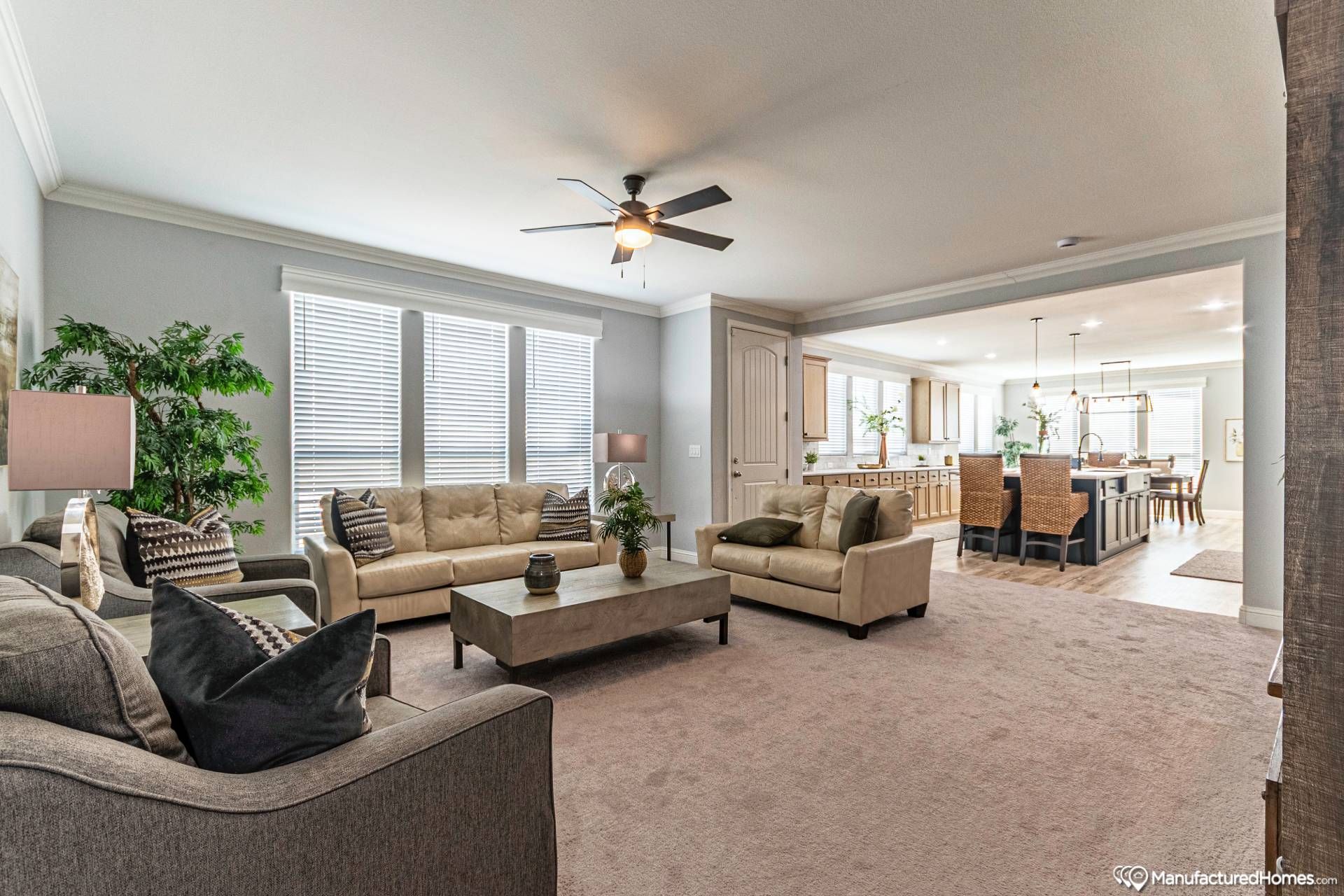 Modern living room with neutral tones, featuring a gray sofa set, coffee table, ceiling fan, large windows with blinds, and an open layout to a dining area.