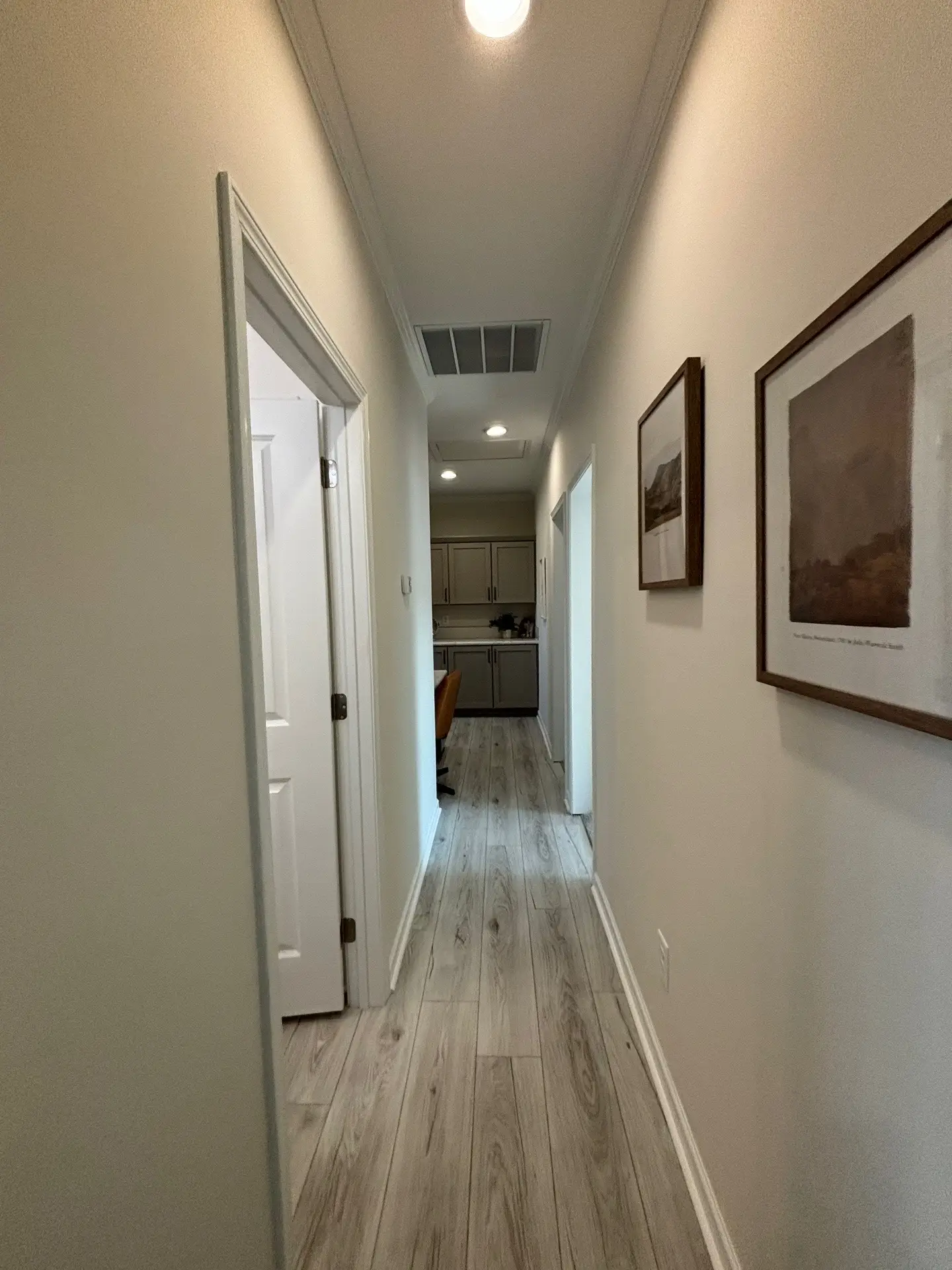 A narrow hallway with light wood flooring leads to a kitchen. Soft, warm lighting and landscape artwork on the wall create a cozy atmosphere.
