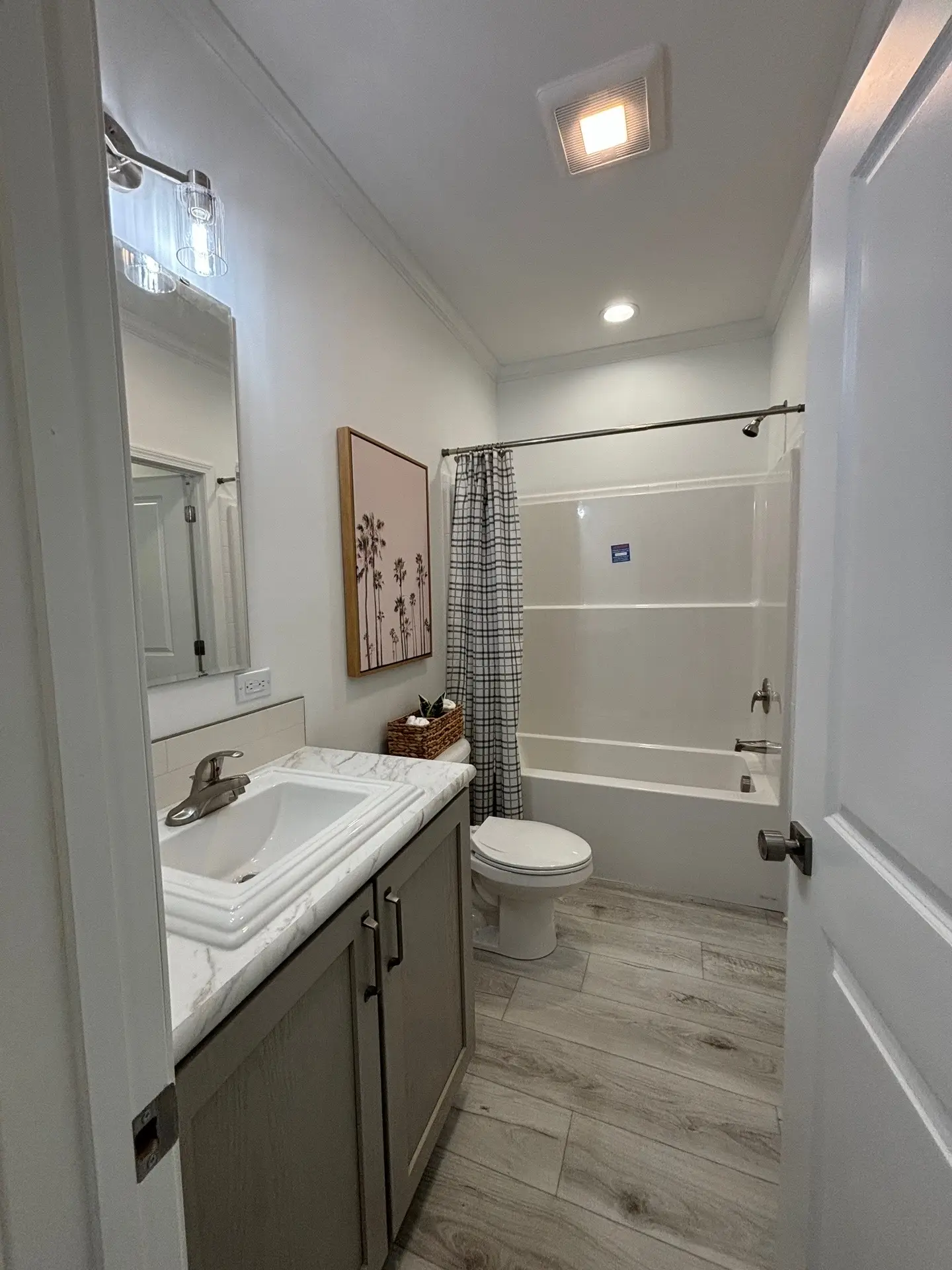 A small, modern bathroom with wood-effect tile flooring. It features a vanity sink with a marble countertop, a toilet, a bathtub with a plaid shower curtain, and a framed wall art above a wicker basket. Bright lighting creates a clean, fresh ambiance.