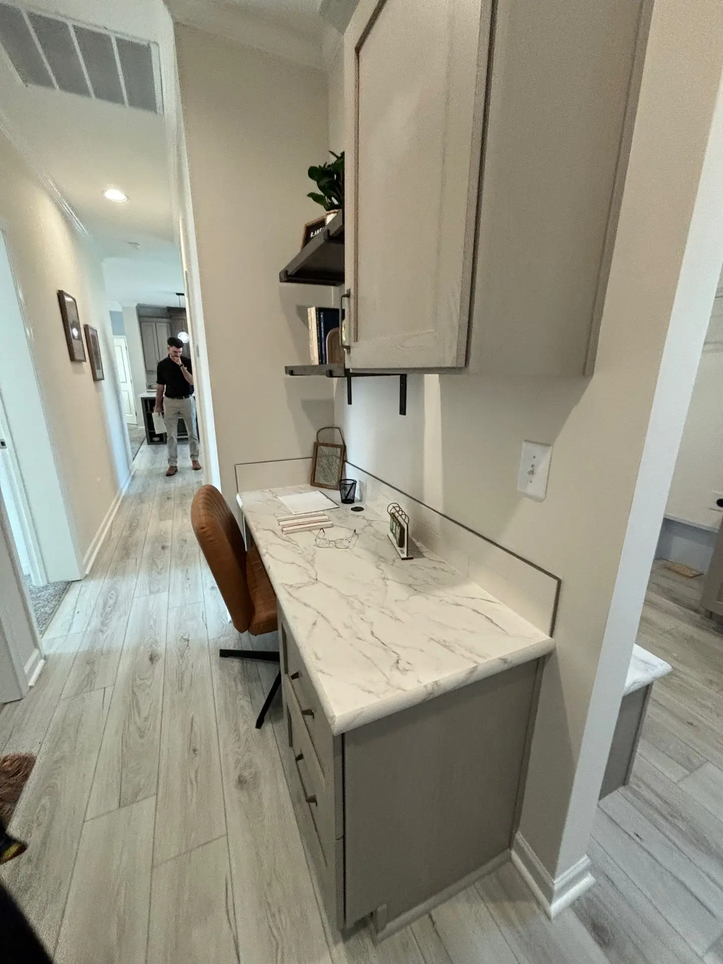 Cozy home office nook with white marble desk, brown leather chair, and overhead shelves. Light wood flooring extends to a distant hallway.