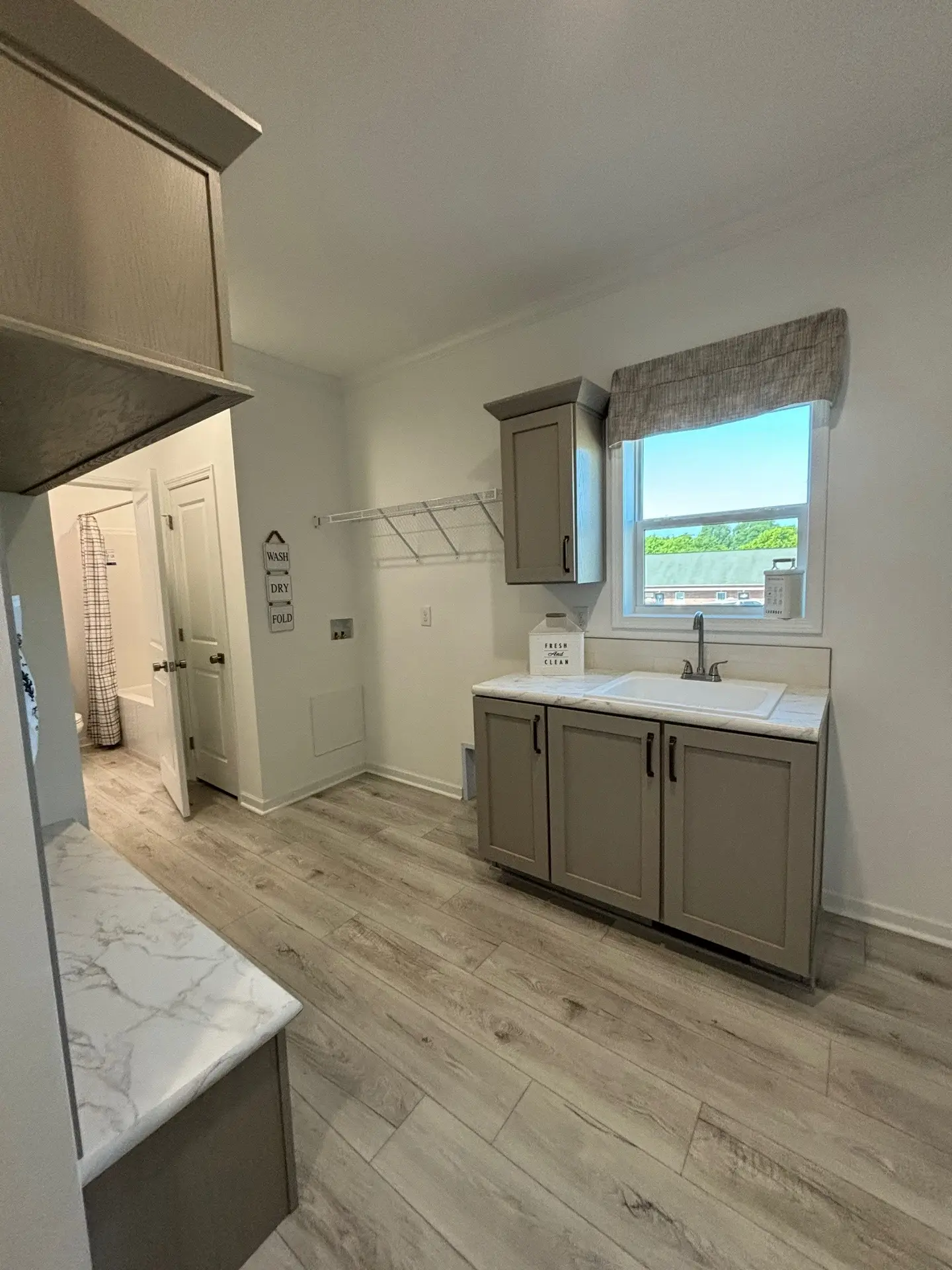 A bright laundry room with light wood flooring and beige cabinets. A window above the sink offers a view of greenery. Wall decor adds a cozy touch.