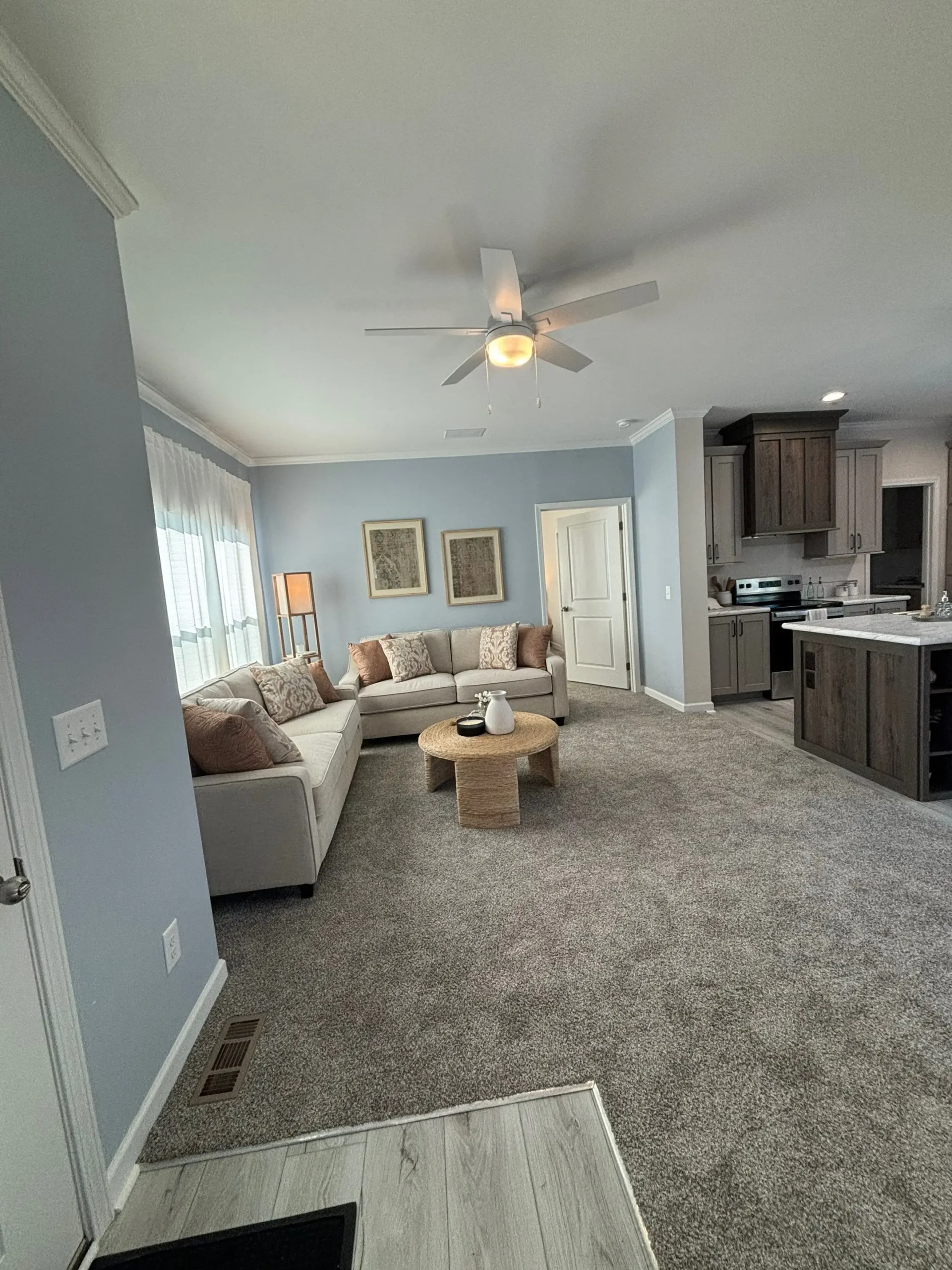 Cozy living room with gray carpet and light blue walls, featuring a beige sectional sofa, wooden coffee table, and ceiling fan. Adjacent kitchen is visible.