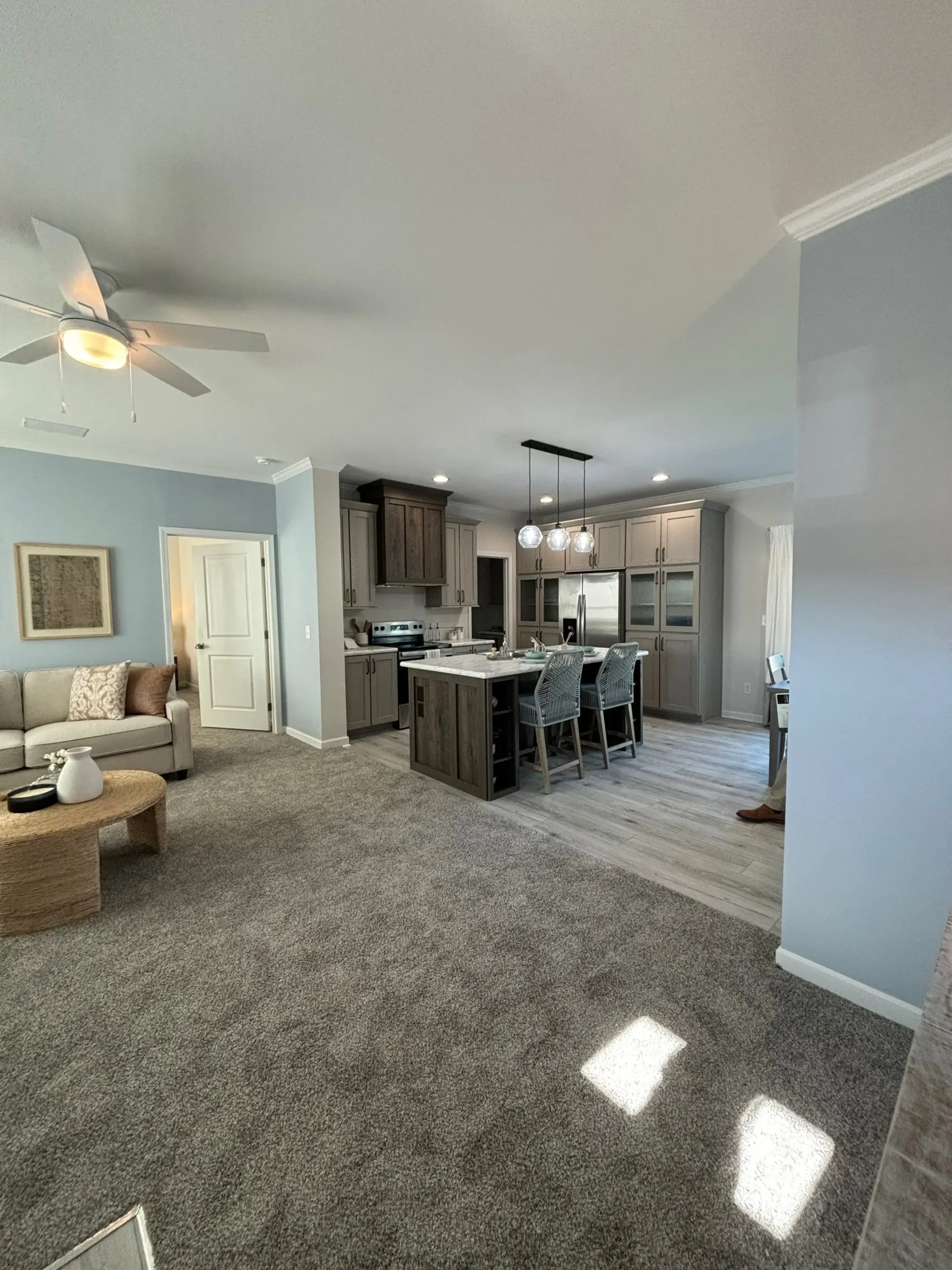 Modern open-concept living area with gray carpet, light walls, ceiling fan, cozy sofa, and kitchen featuring an island, bar stools, and pendant lights.