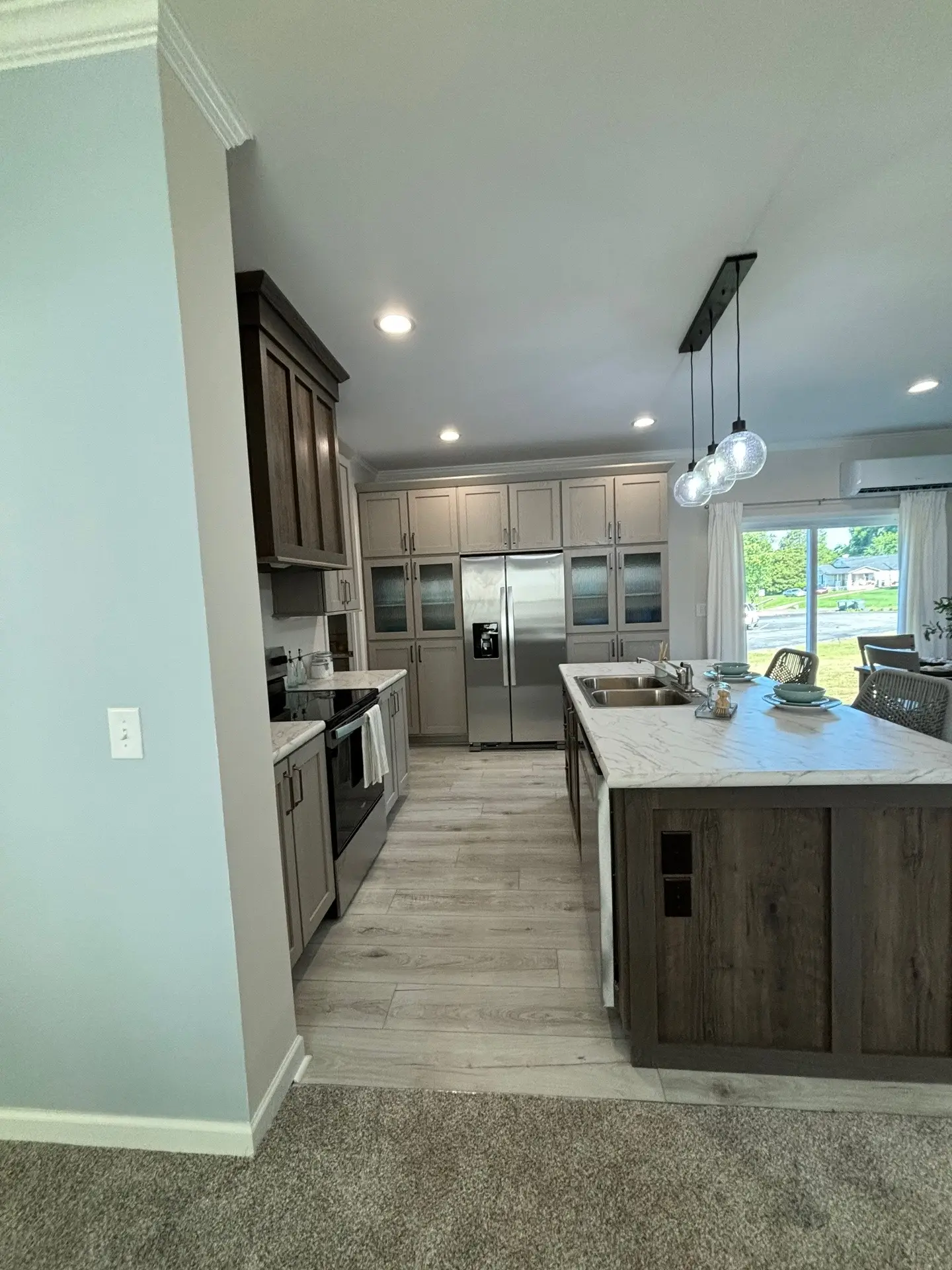 Modern kitchen with wood cabinets, stainless steel appliances, and marble countertops. An island with pendant lights and a nearby window creates an airy feel.