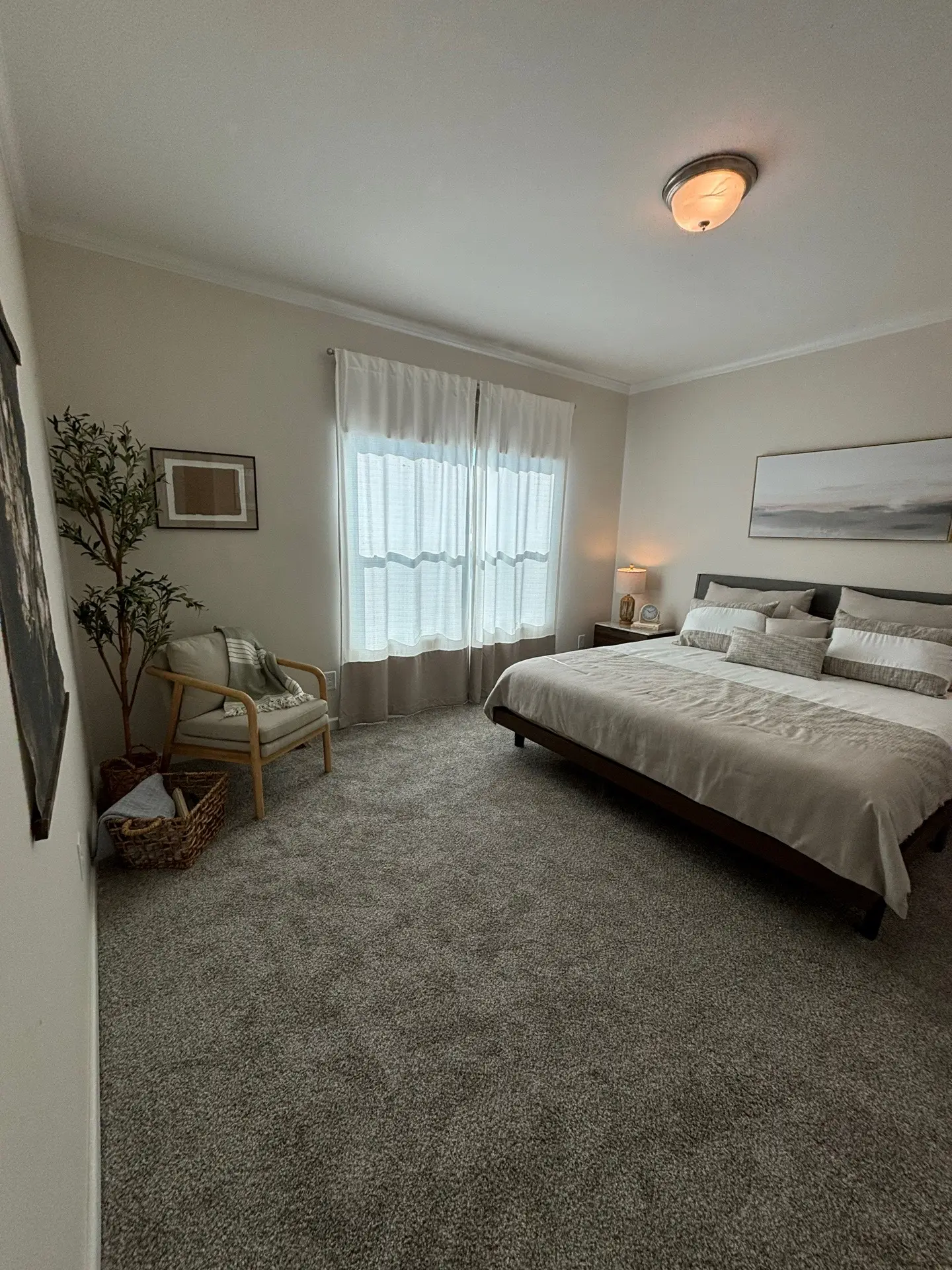 A cozy bedroom with a large bed featuring white and gray bedding. Soft curtains filter light from a window. A chair and a plant add warmth and tranquility.
