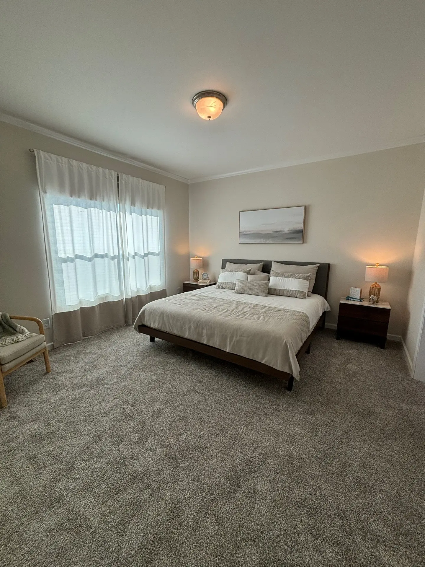 A serene bedroom with a large bed, beige bedding, and multiple pillows. Soft lighting from table lamps, sheer curtains, and a simple wall art create a calm ambiance.