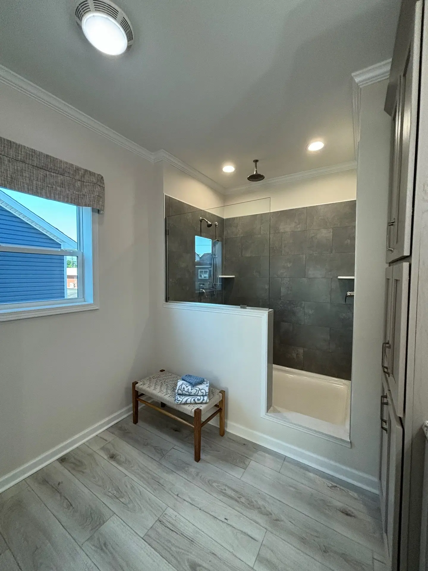 Modern bathroom with gray tile shower, glass door, and overhead light. A wooden bench with folded towels sits on light wood flooring. Airy and clean.