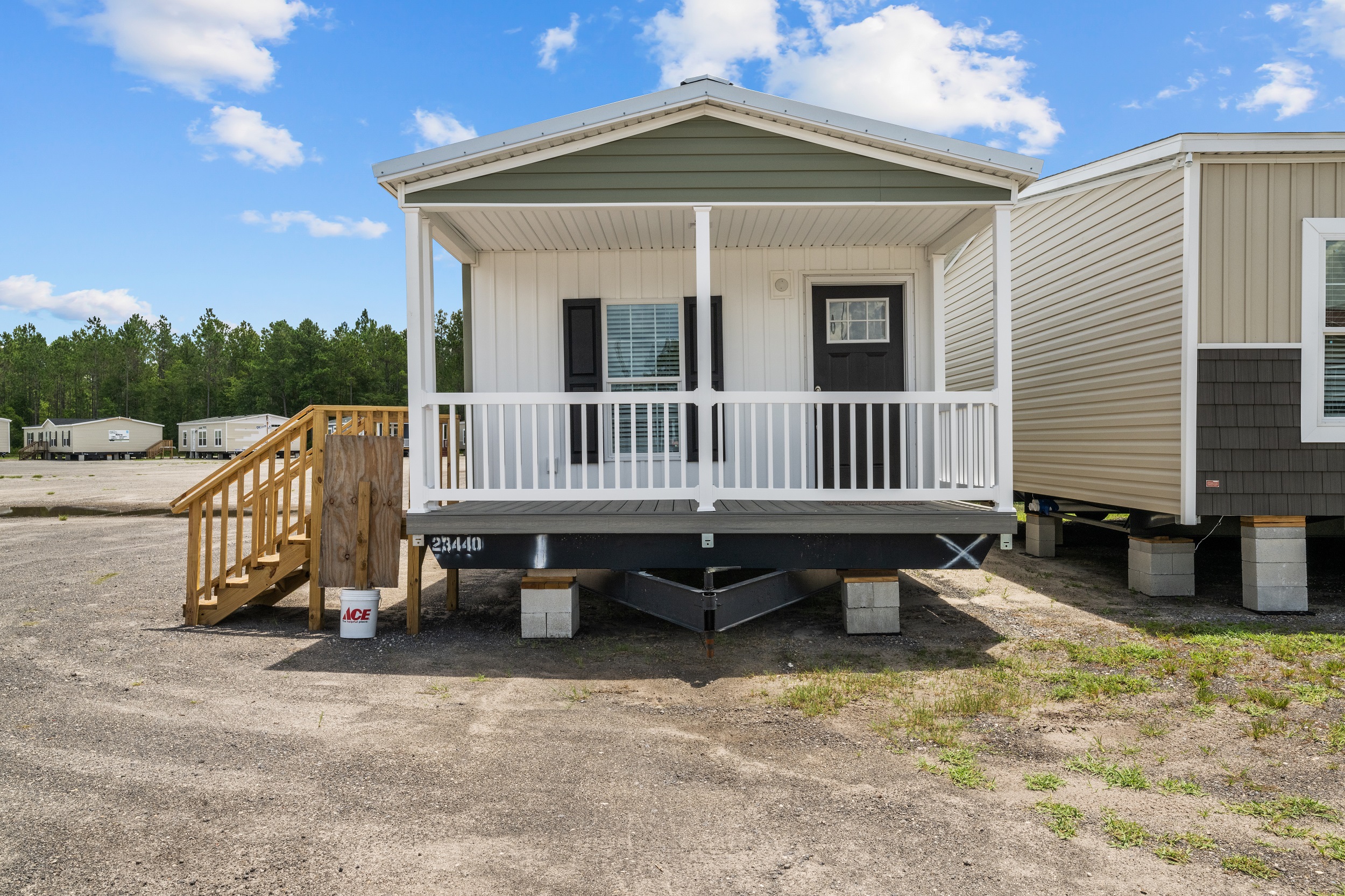 Compact mobile home with a covered porch and wooden steps, set on a gravel lot. Blue sky with clouds above, surrounded by trees in the distance.