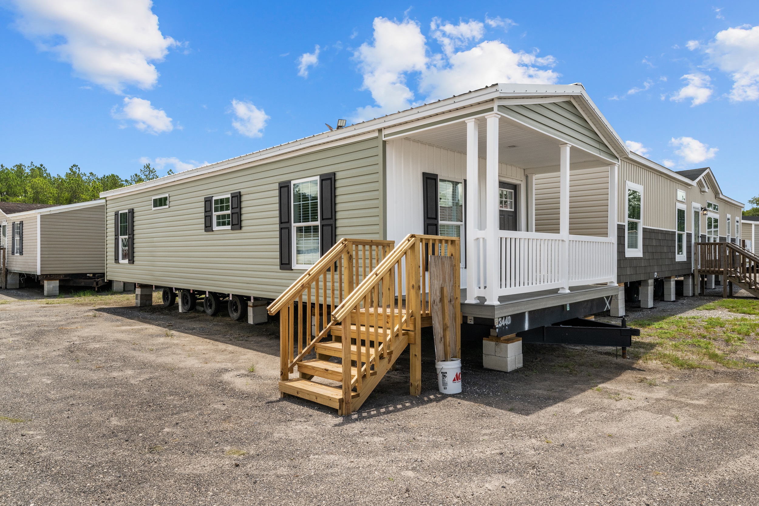 Row of mobile homes under a blue sky with scattered clouds. Each home has a small porch and wooden steps, conveying a clean and welcoming atmosphere.