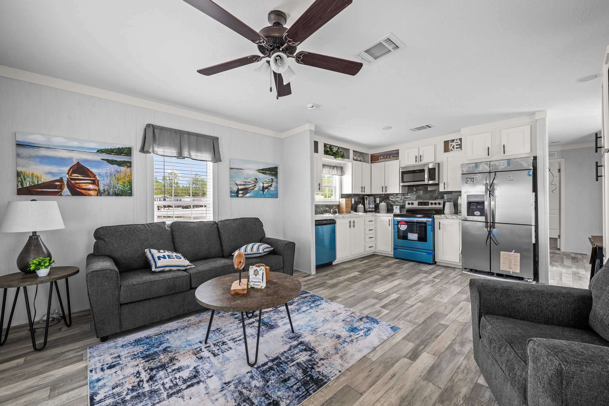 Modern living room with gray sofas, a round wooden coffee table, and a blue-accented kitchen with white cabinets. Artwork depicts serene boats.