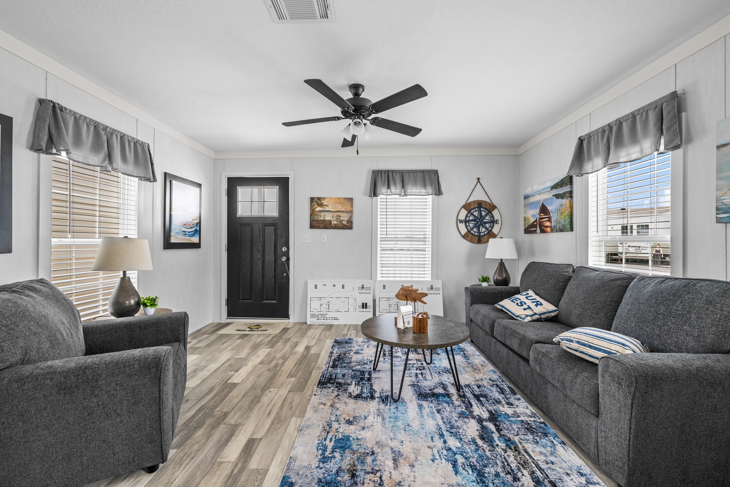 Stylish living room with gray sofas, a round coffee table, and a blue patterned rug. Wall art, a ceiling fan, and natural light create a cozy atmosphere.
