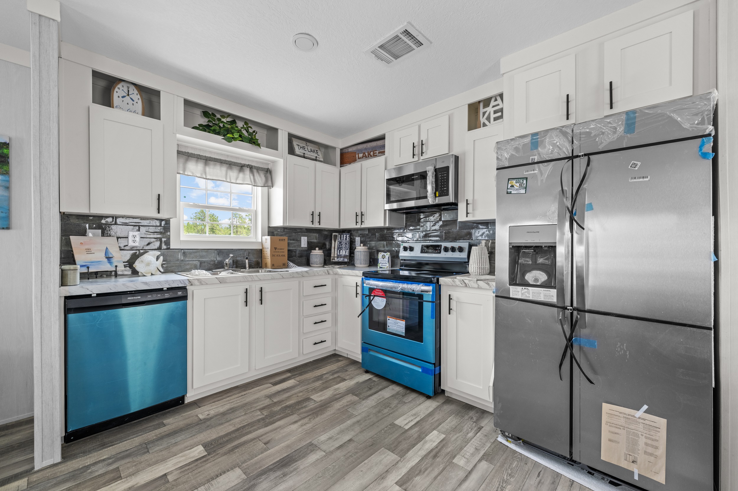 Modern kitchen with white cabinets, stainless steel appliances, and light gray wood flooring. A bright window and decorative elements add a fresh feel.