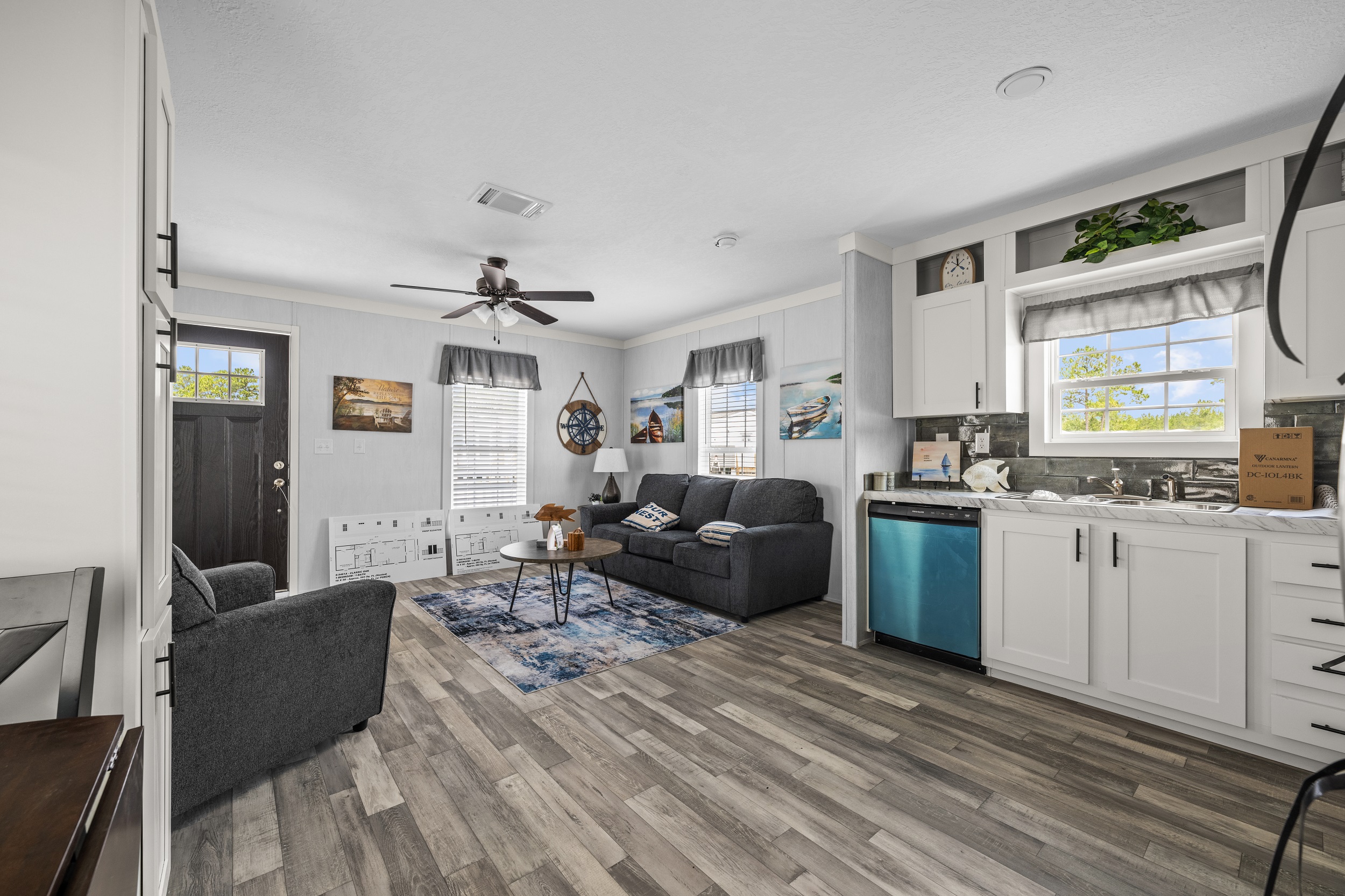 Cozy living room with gray couches, a blue patterned rug, and a small coffee table. Bright kitchen with white cabinets and a teal dishwasher. Modern decor.