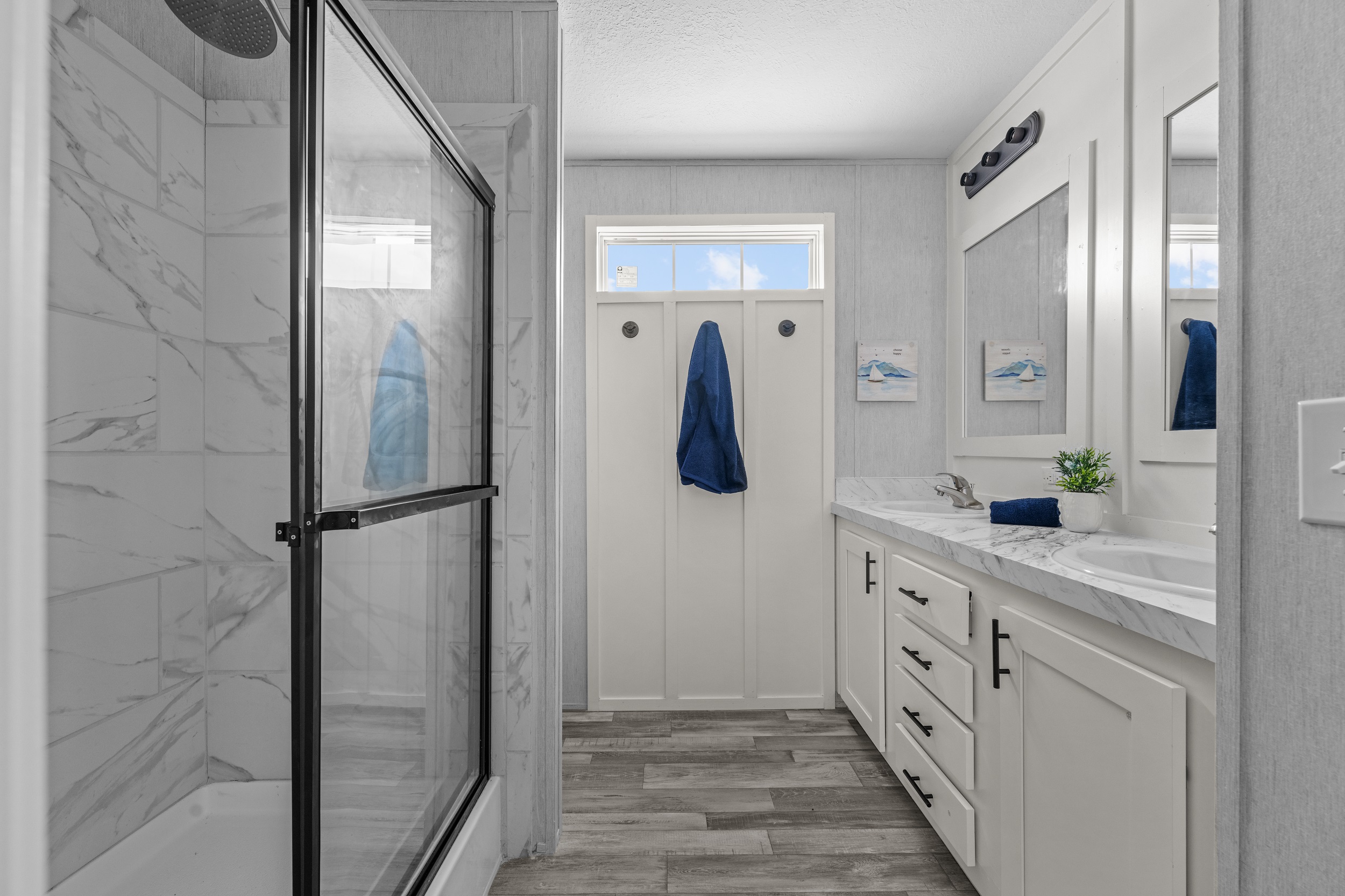 Modern bathroom with a marble-tiled shower and glass door on the left. White vanity with dual sinks and marble countertop on the right, navy accents, and wall art.