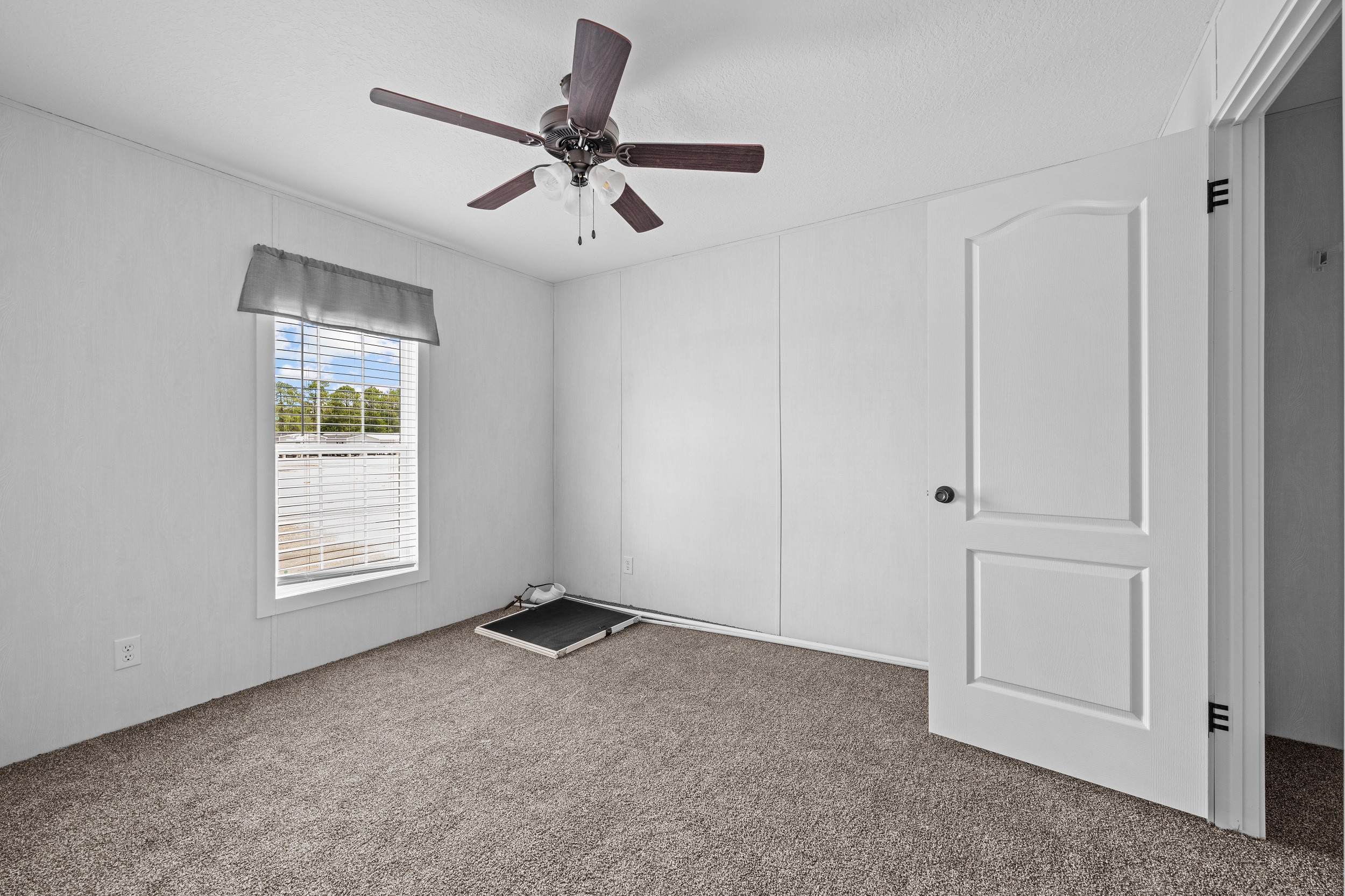 Empty room with beige carpet, white walls, and a ceiling fan. A window with blinds and a valance lets in light, showing a calm, neutral tone. An open door is visible.