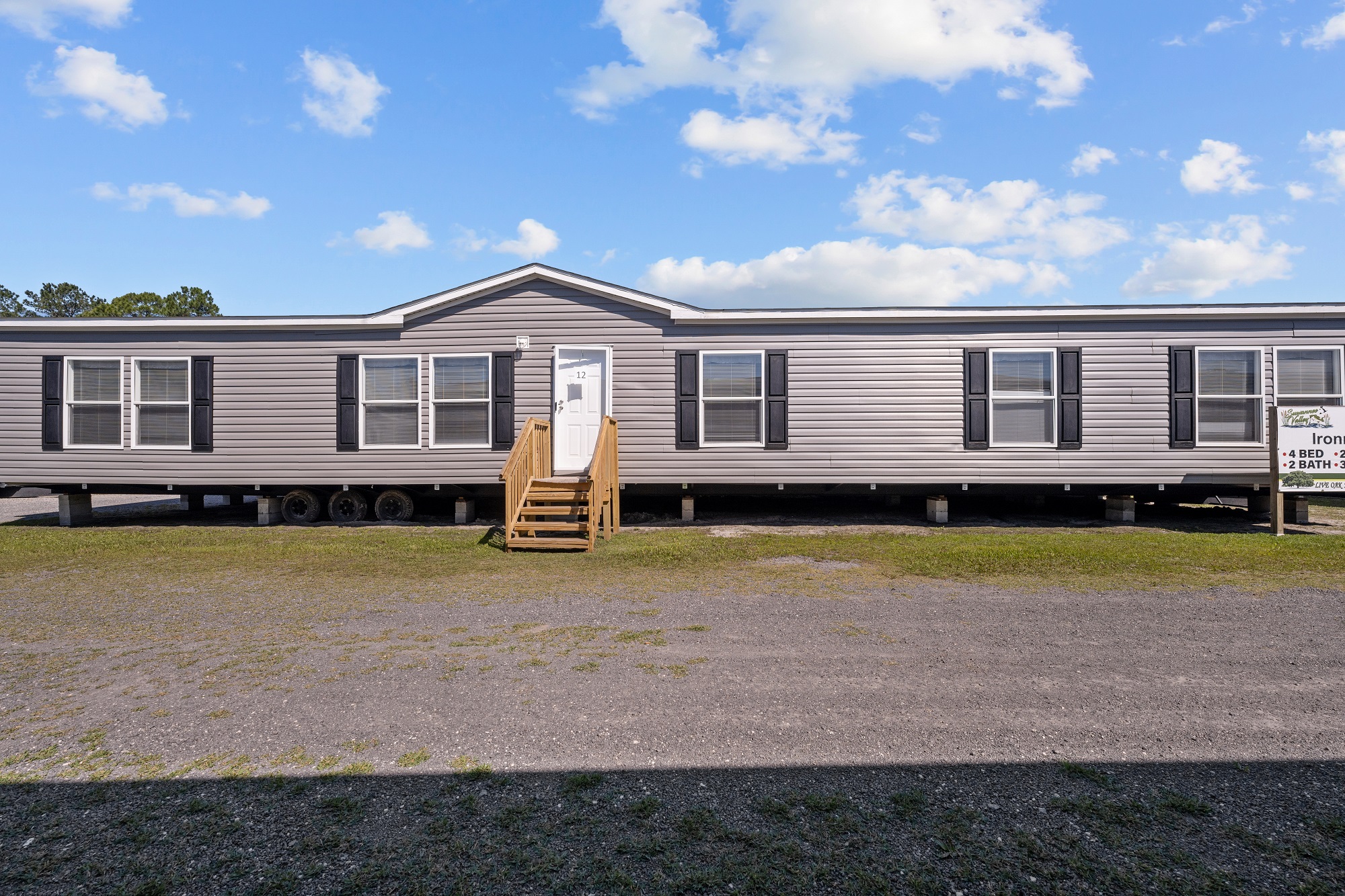 A beige, single-story mobile home with black shutters on raised ground, fronted by a small wooden staircase. The sky is clear and blue, creating a calm atmosphere.