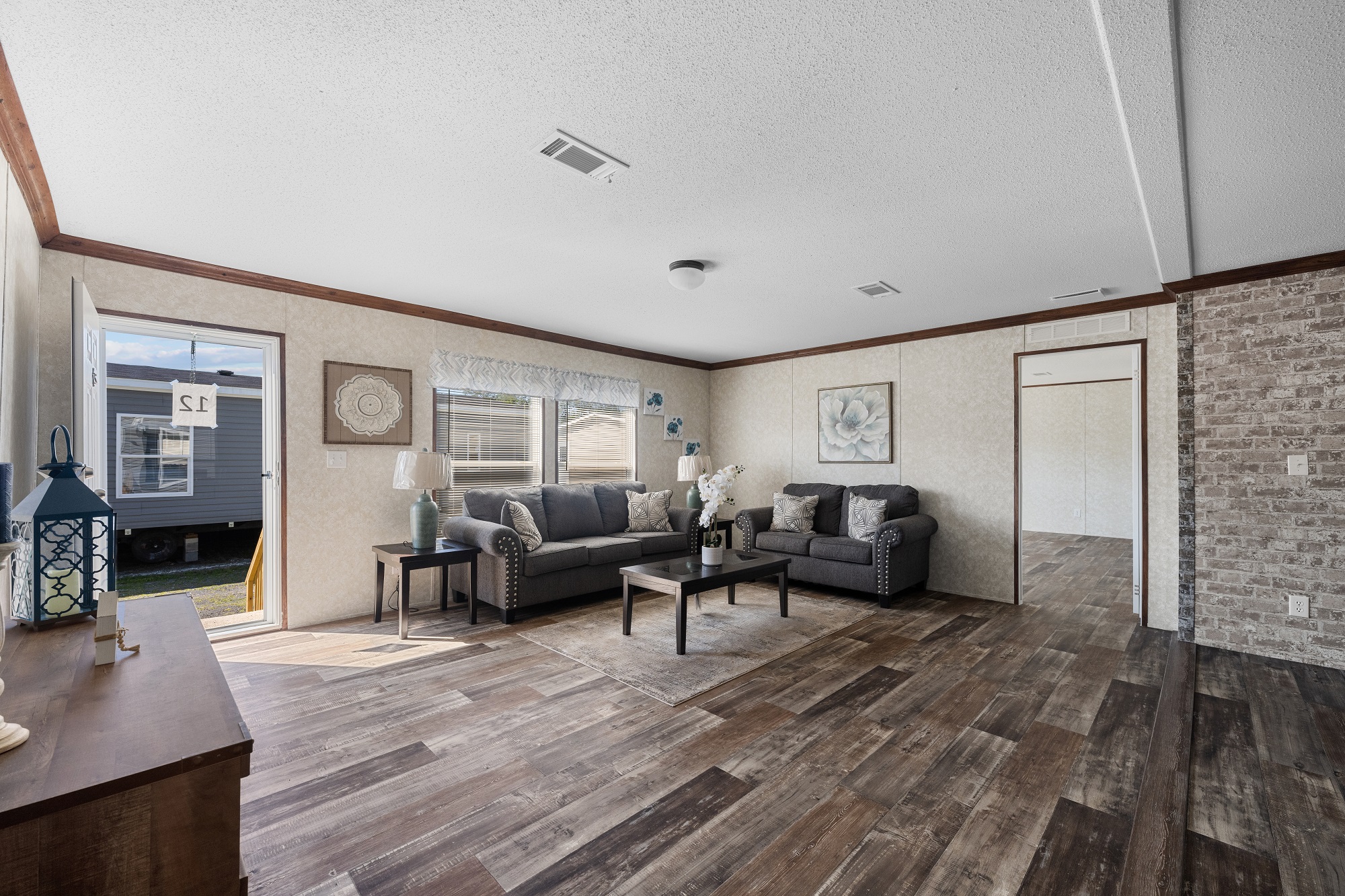 Spacious living room with two gray sofas, a wooden coffee table, and decorative plants. Natural light from large window, wood floor, welcoming tone.
