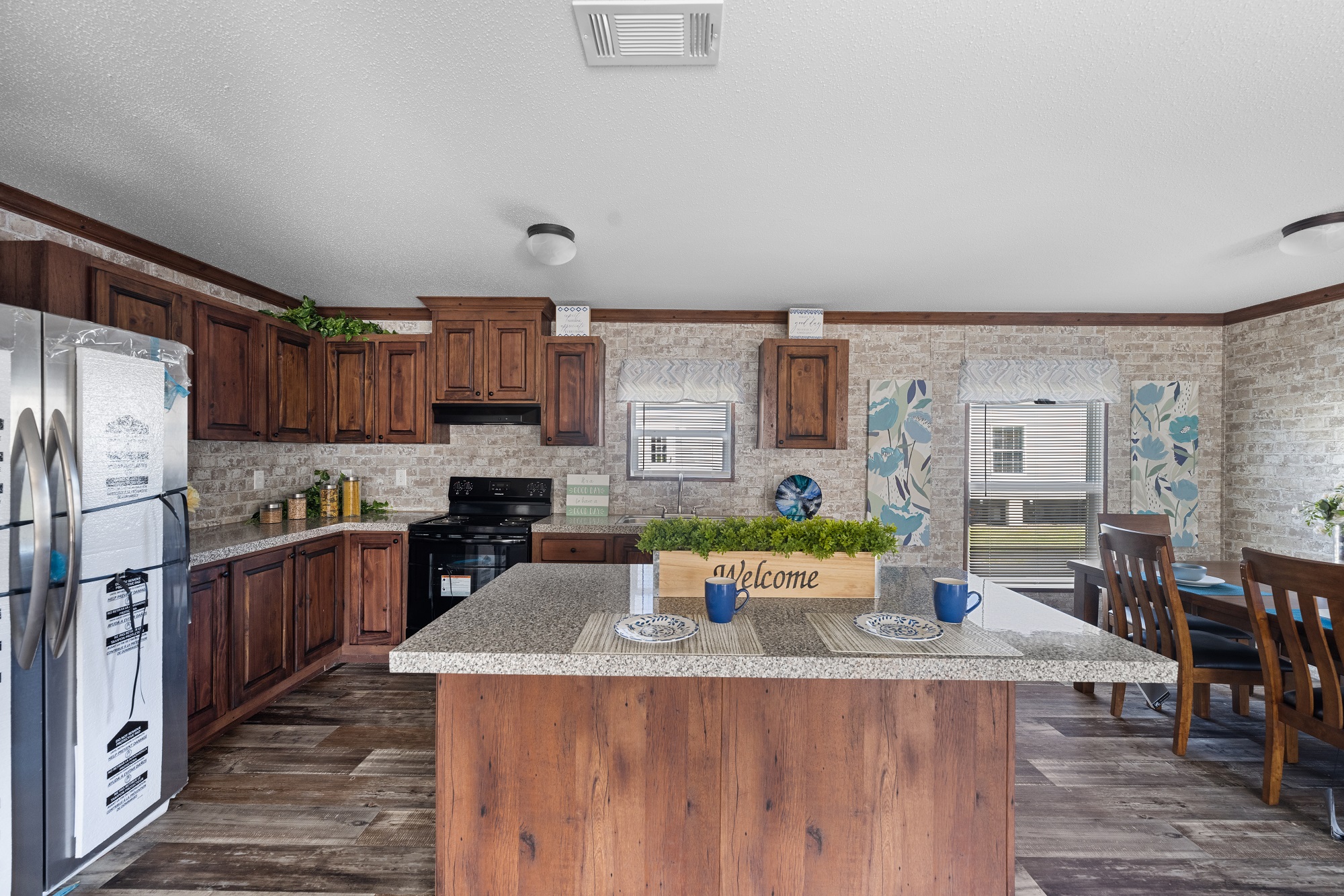 A cozy kitchen with wooden cabinets, a central island with a "Welcome" sign, blue cups, and placemats. Light decor and wood accents create a warm, inviting atmosphere.