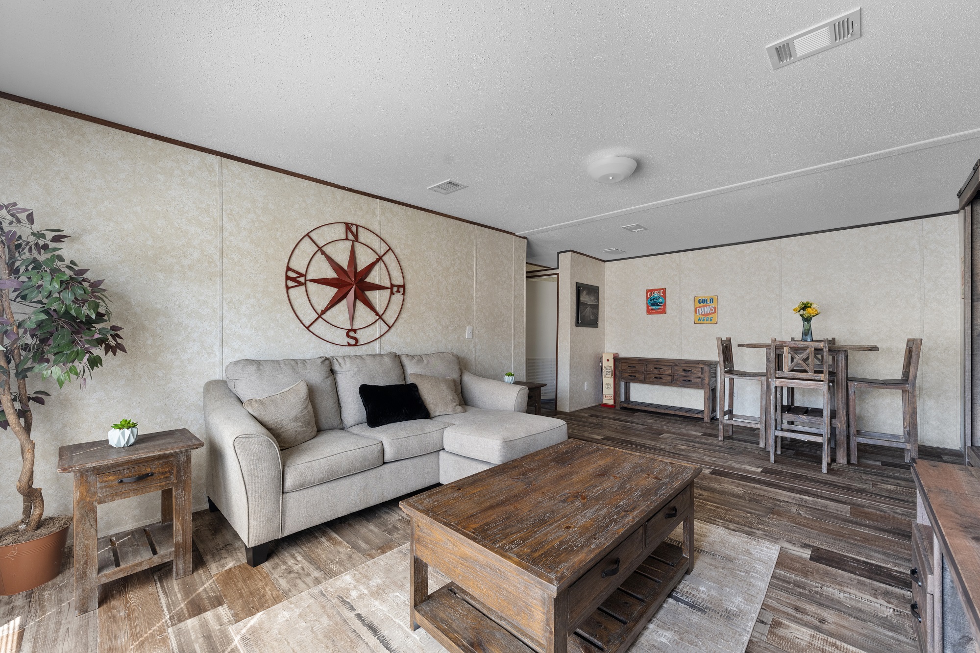 A cozy living room with a beige sectional sofa and decorative compass wall art. The space features wooden furniture, a rug, and a dining area with flowers.
