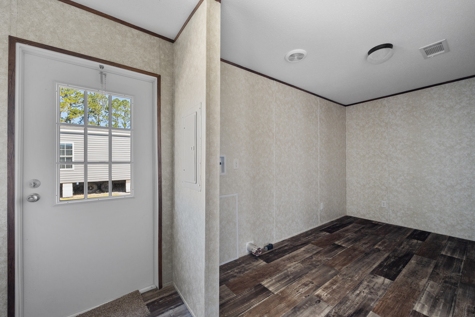A small, empty room with plain beige walls and dark wooden floors. A white door with a window shows an outside view of trees. The space feels minimal and simple.