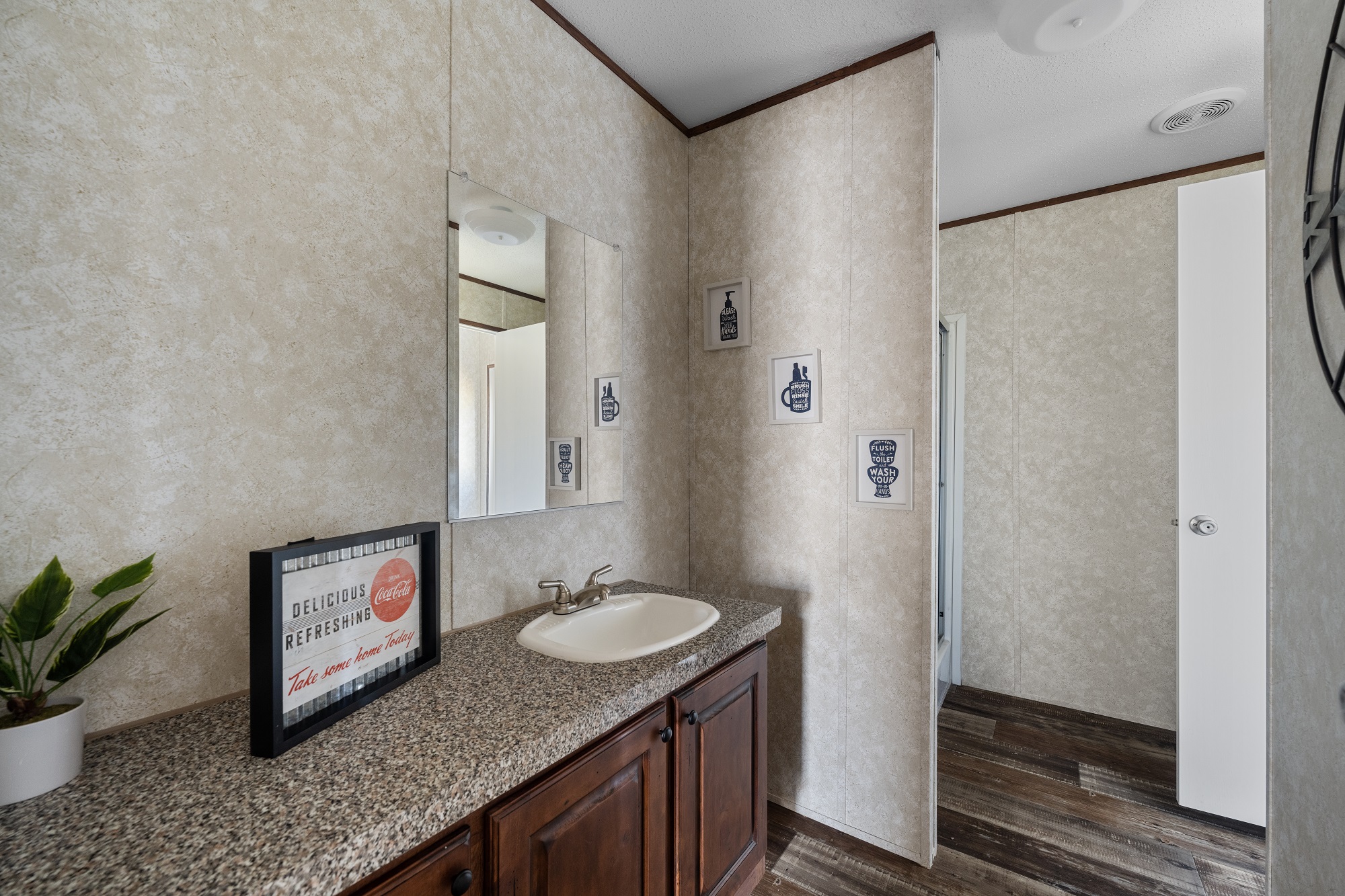 A bathroom with a granite countertop, a white sink, and wooden cabinets. A mirror is above the sink, and minimalist wall art decorates the light-toned walls. A small plant and framed artwork add a cozy touch.
