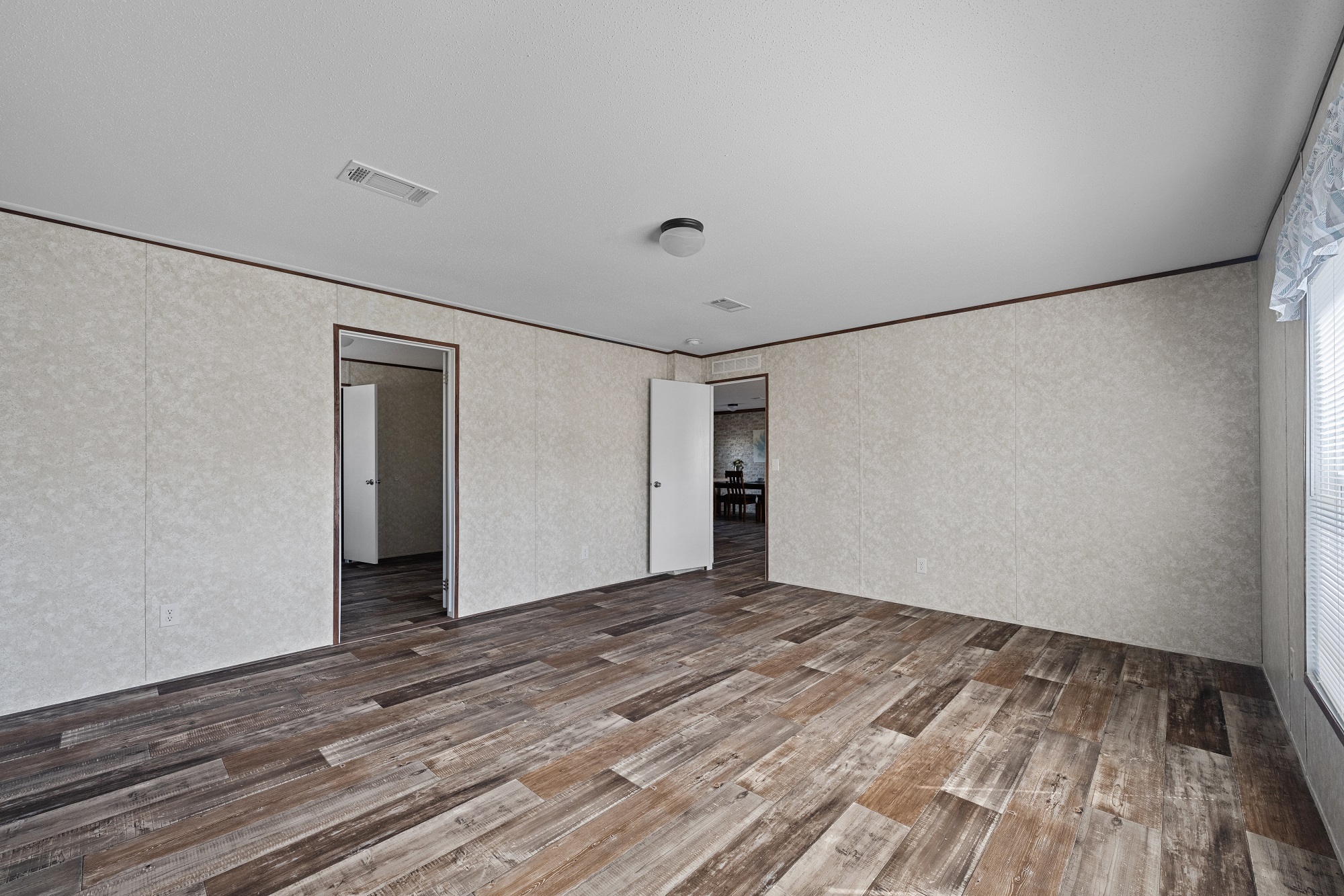 Empty room with wooden flooring, light beige walls, and white ceiling. Two open doors lead to adjacent rooms. A window on the right lets in natural light.