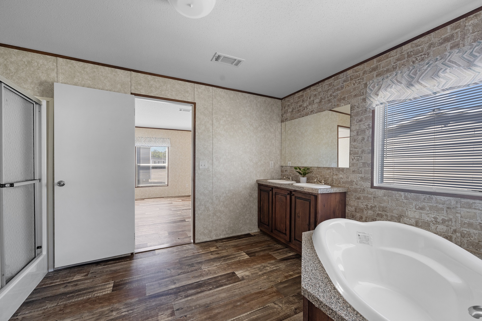 Spacious bathroom with a large bathtub, wooden cabinets, and a brick-patterned wall. A shower and doorway lead to a bright room with wooden flooring.