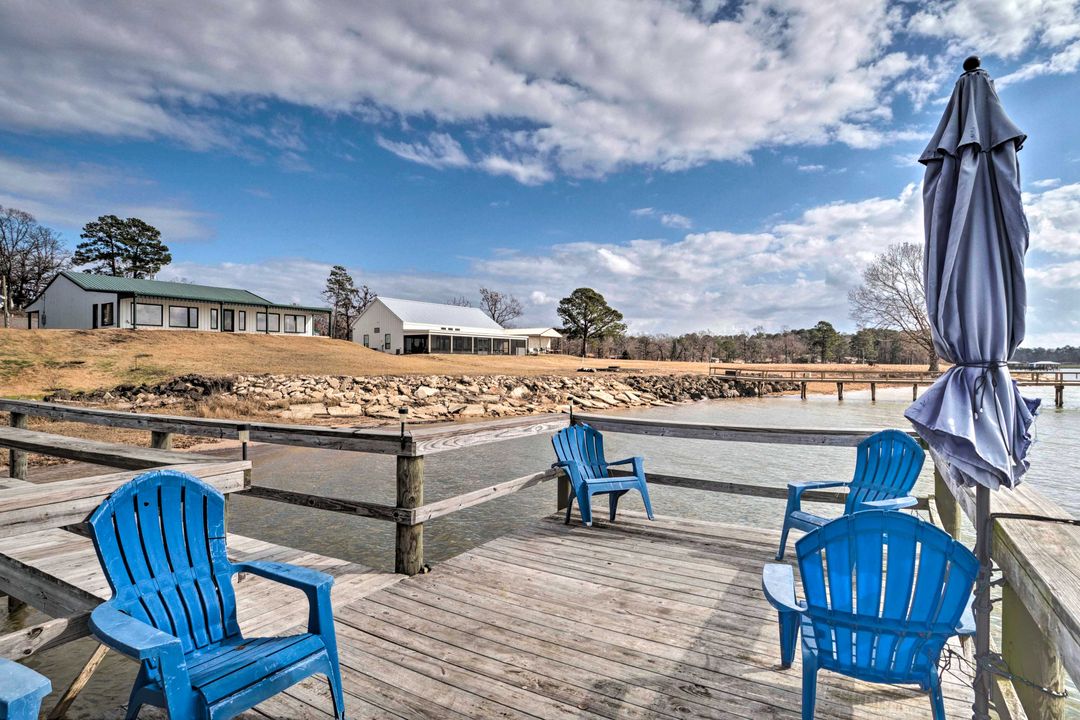 Wooden dock with blue chairs and a closed umbrella overlooks a serene lake. In the background, houses and trees are under a partly cloudy sky. Peaceful atmosphere.