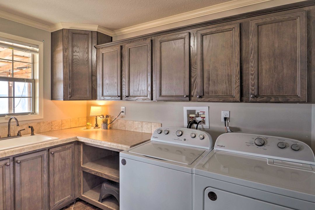 A cozy laundry room with wooden cabinets, a countertop, and a sink. A washing machine and dryer are side by side with warm lighting above.