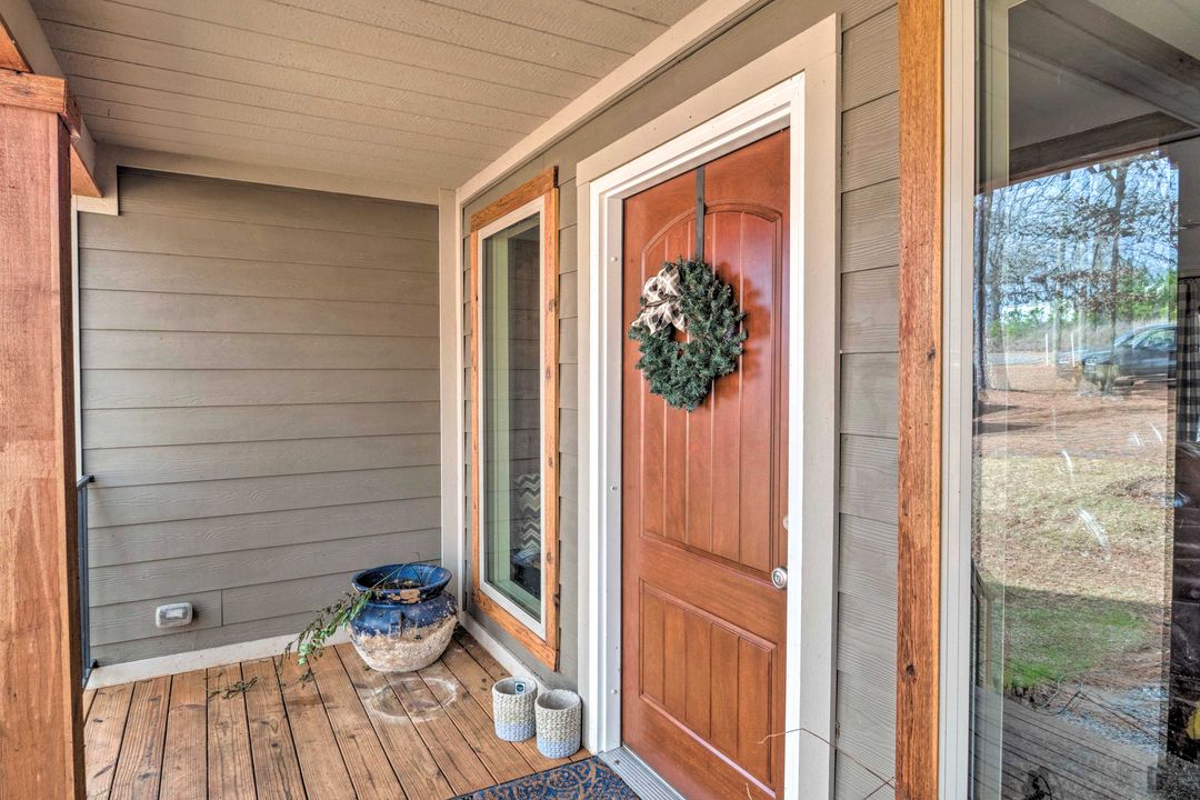 Front porch with wood paneling, featuring a brown door adorned with a green wreath. A blue pot and woven baskets sit on the wooden floor. A window reflects trees outside.