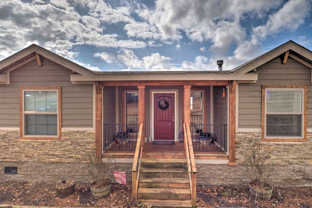 Single-story house with wooden accents, surrounded by tall pine trees, beside a large garage. An American flag waves in the breeze under a partly cloudy sky.